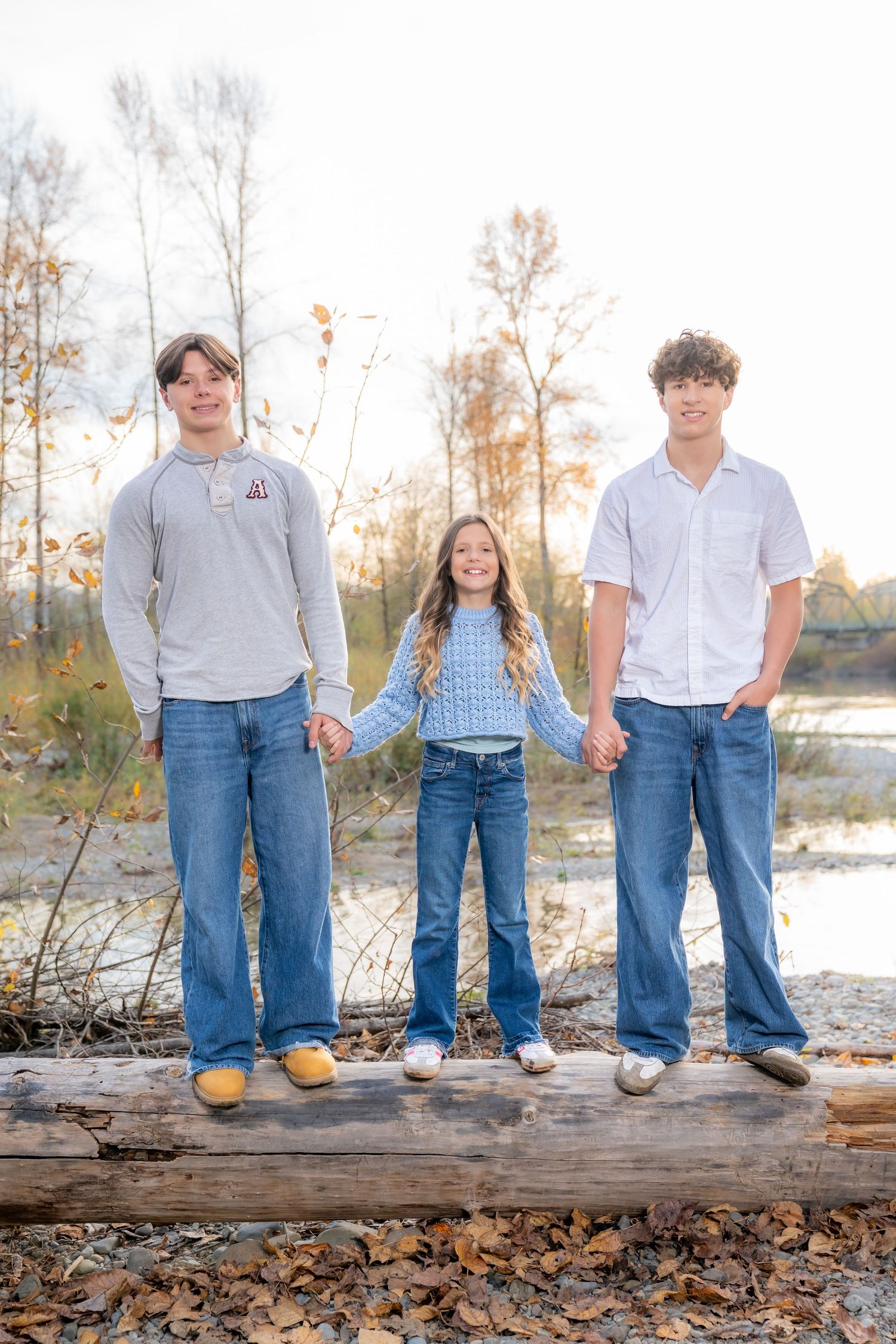 Group standing on a fallen log by the river during autumn with bare trees in background.