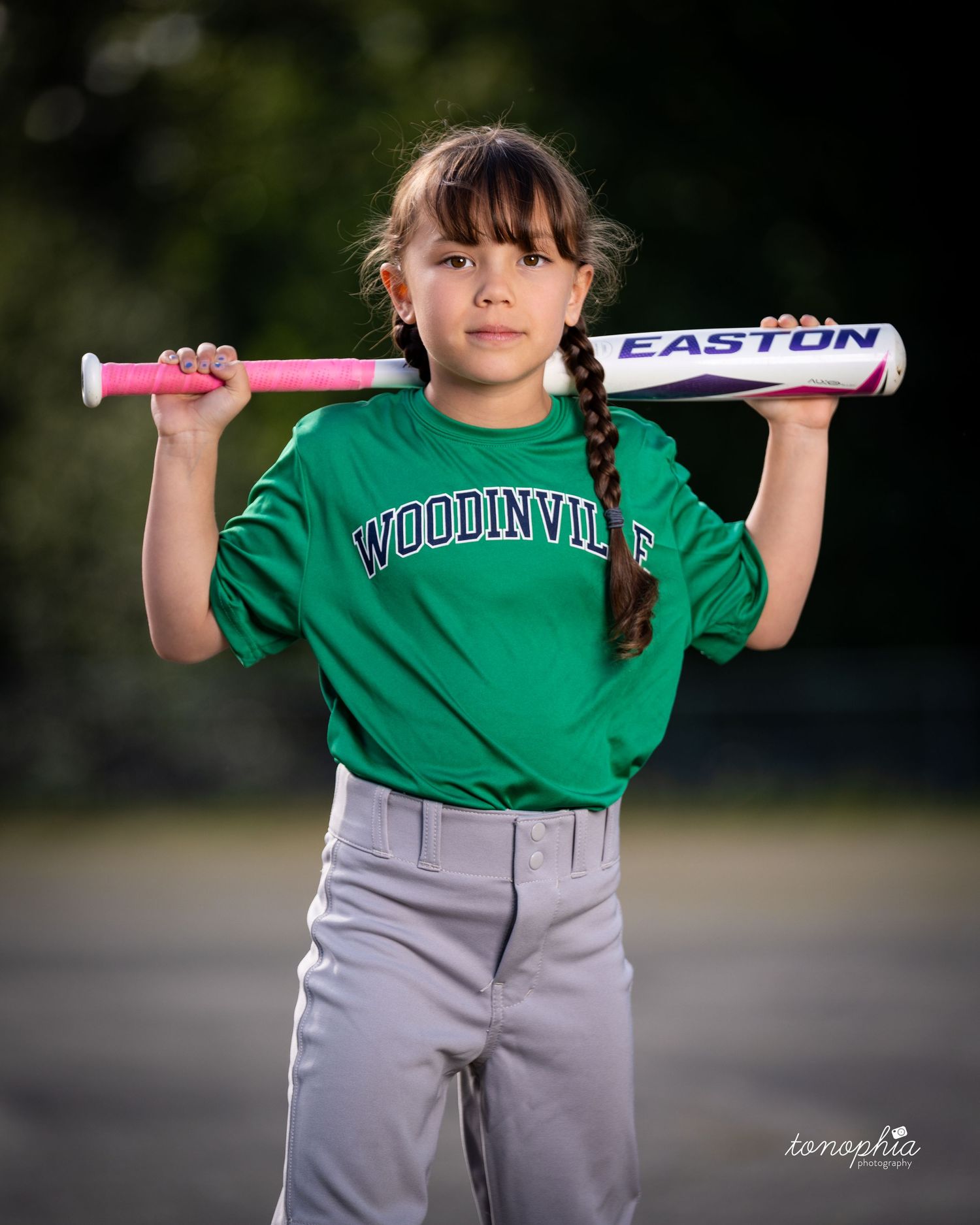 Youth baseball player in green jersey and gray pants holds pink bat in batting stance against blurred outdoor background.