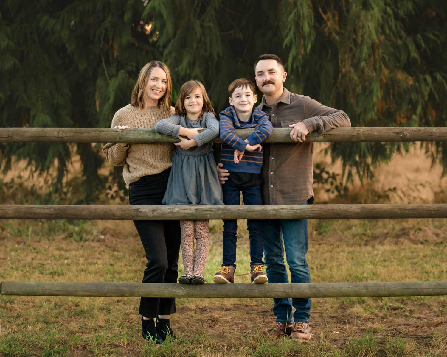 Family group leaning on wooden fence in rustic outdoor setting with evergreen trees.