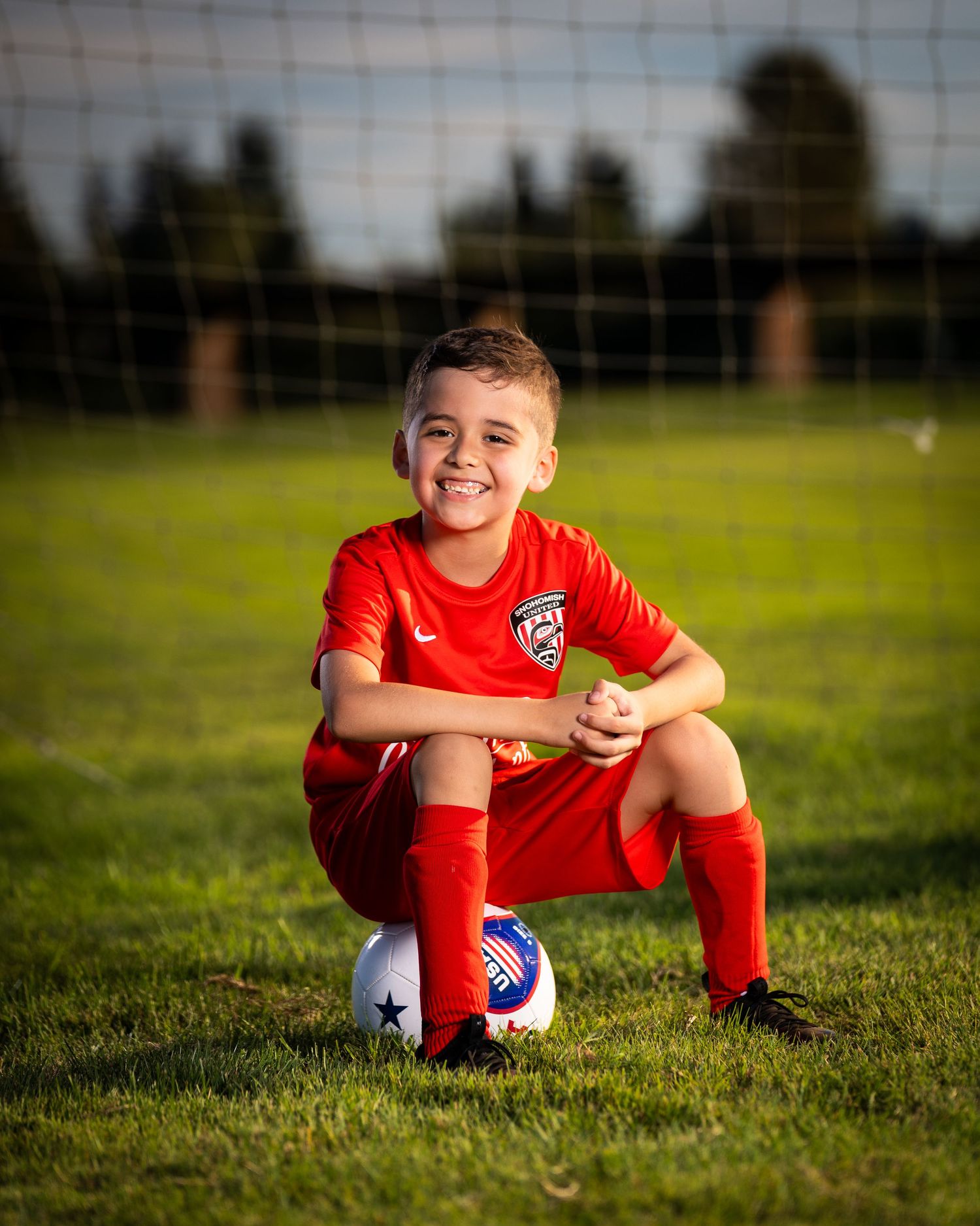 Young soccer player in red uniform sits on grass field with ball and playful expression during practice session.