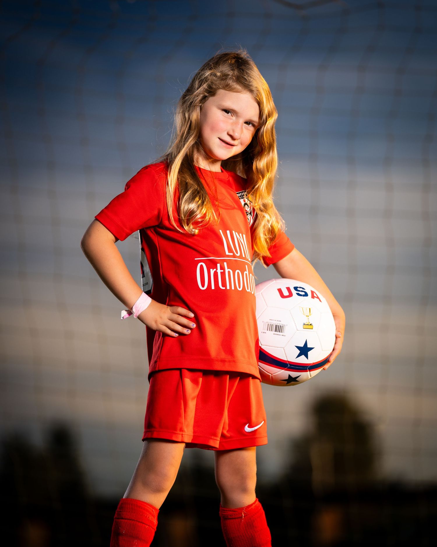 Soccer player in red uniform poses with white ball against dramatic sunset sky.