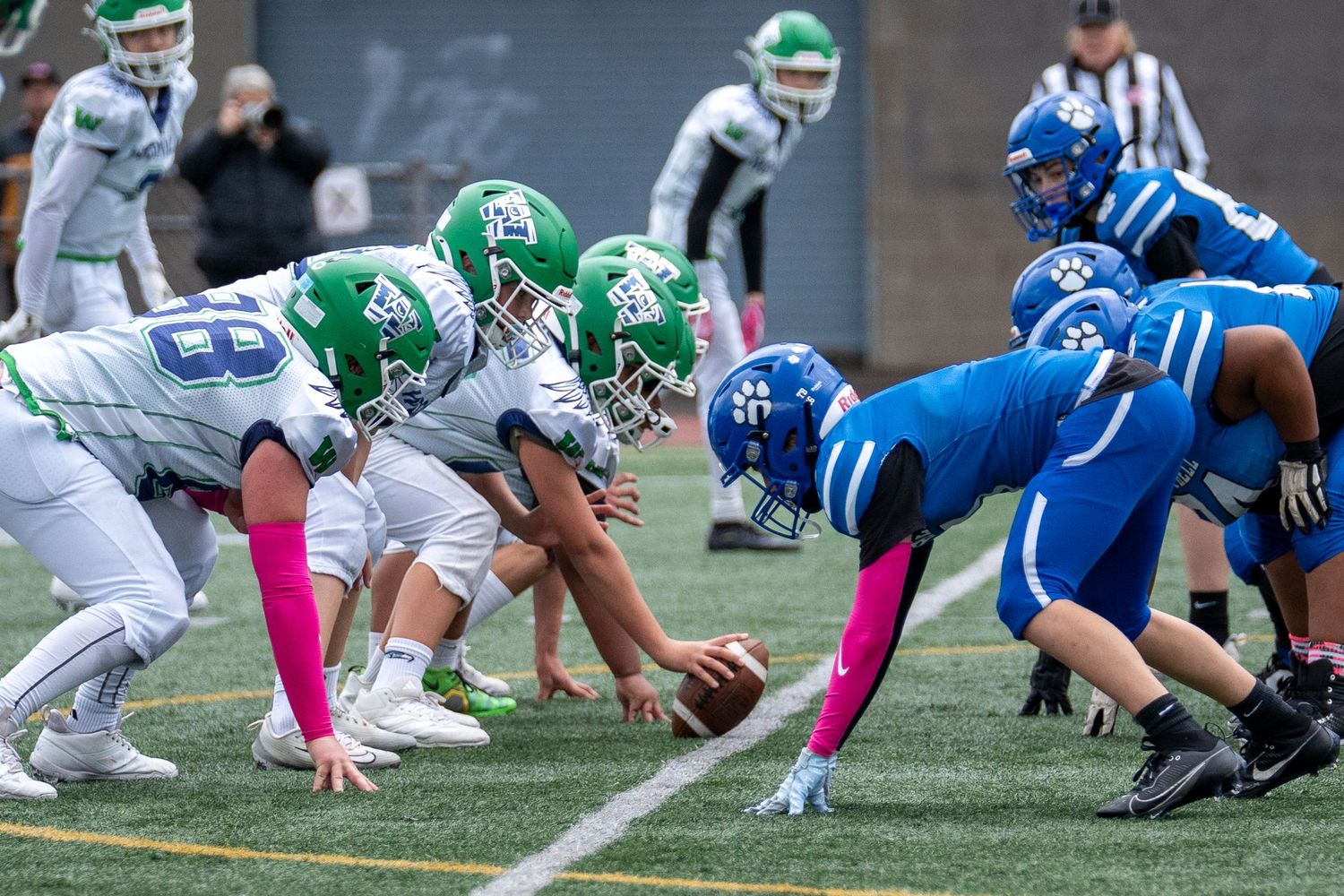 Football teams in blue and green uniforms line up at scrimmage with pink accents for breast cancer awareness.
