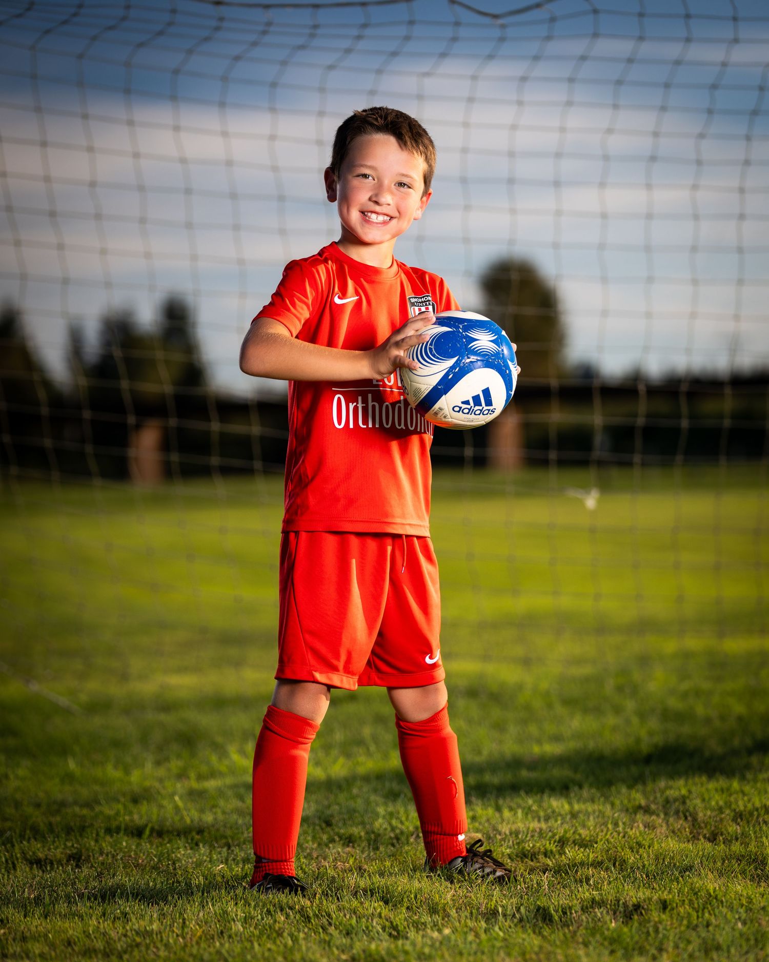 Young soccer player in red uniform poses with a white ball against a soccer net on a sunny field.