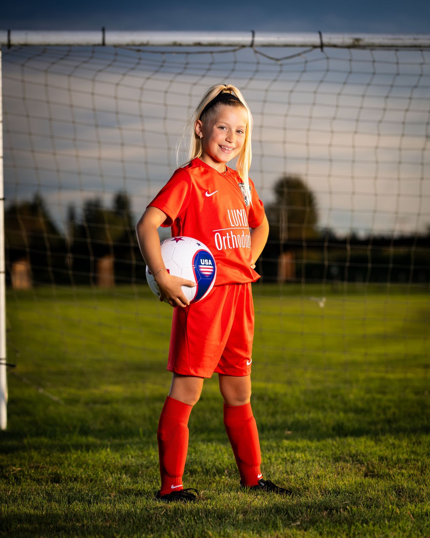Youth soccer player in bright orange uniform poses with ball in front of goal net on green field at sunset.