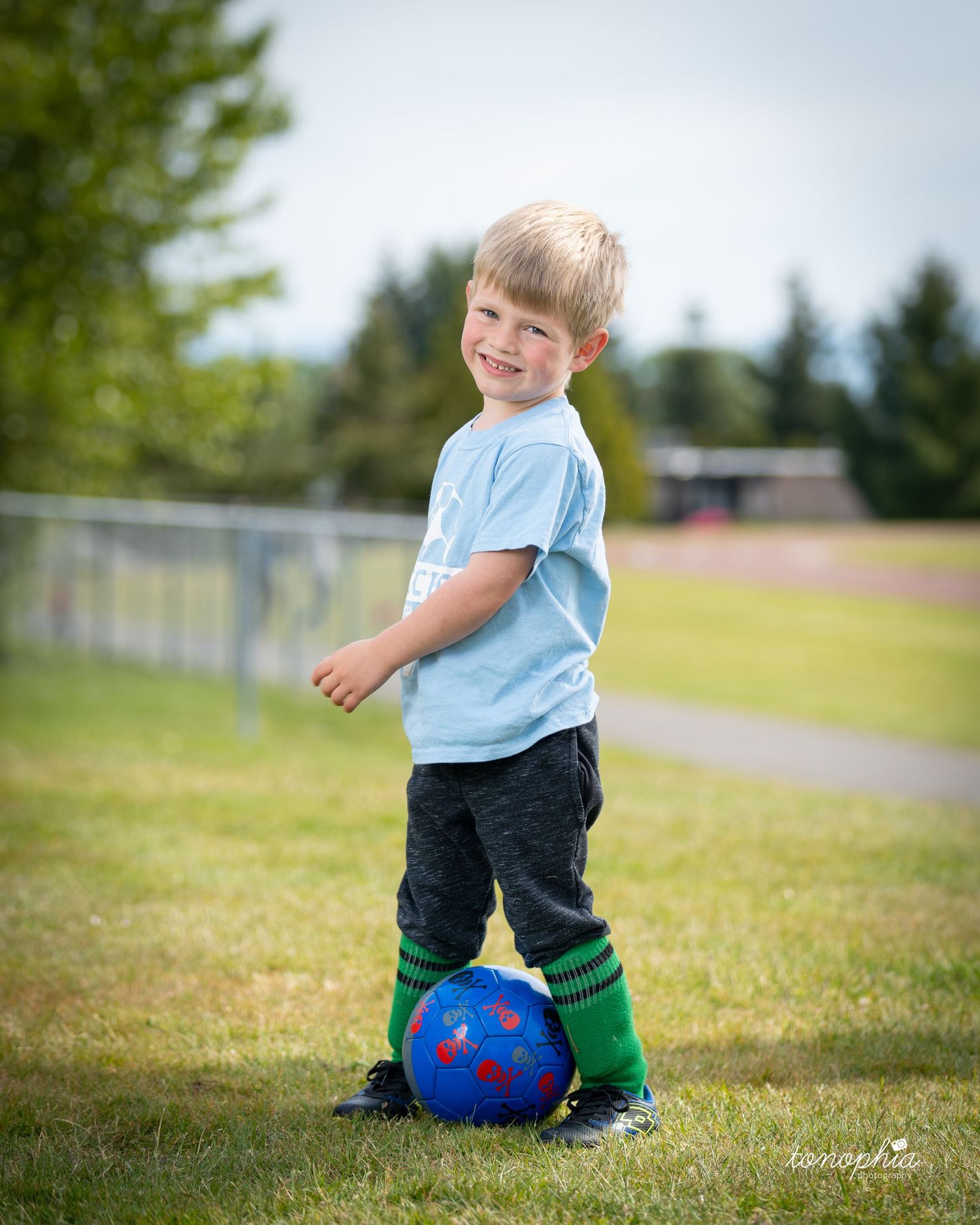 A young child sits on a grassy field with a blue and red soccer ball on a sunny day.