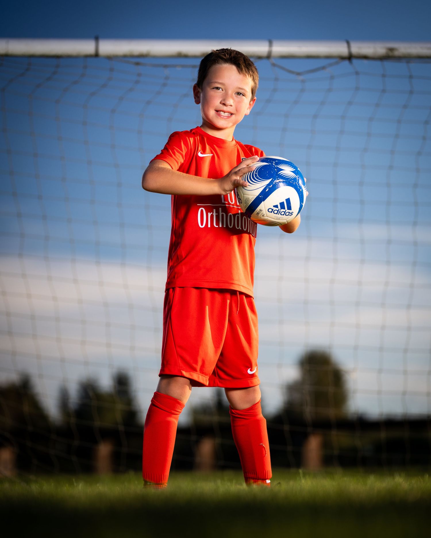 Young soccer player in bright orange uniform poses confidently with ball against soccer goal net at sunset.