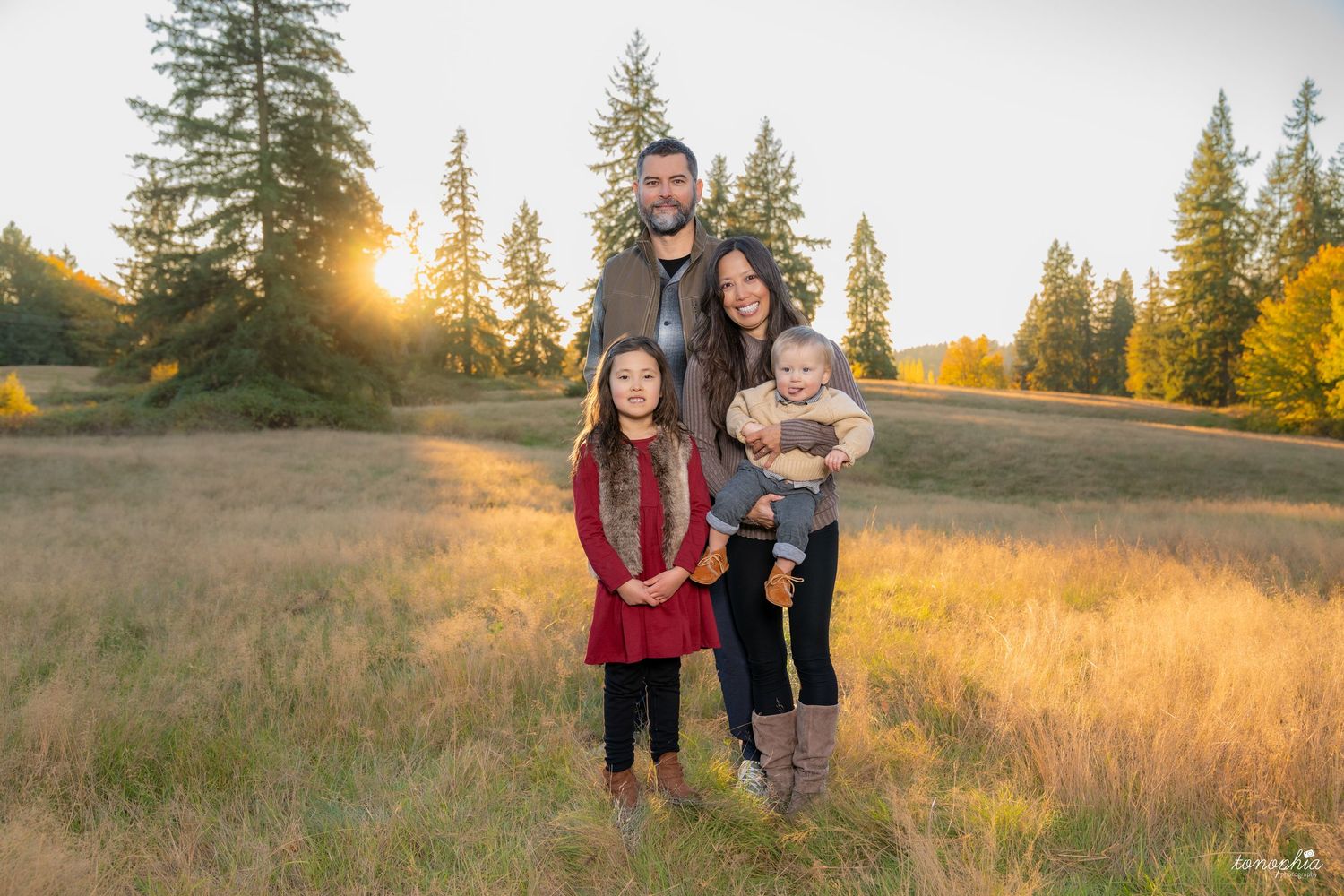 Family enjoying golden hour in a tall grass field with evergreen trees in background.