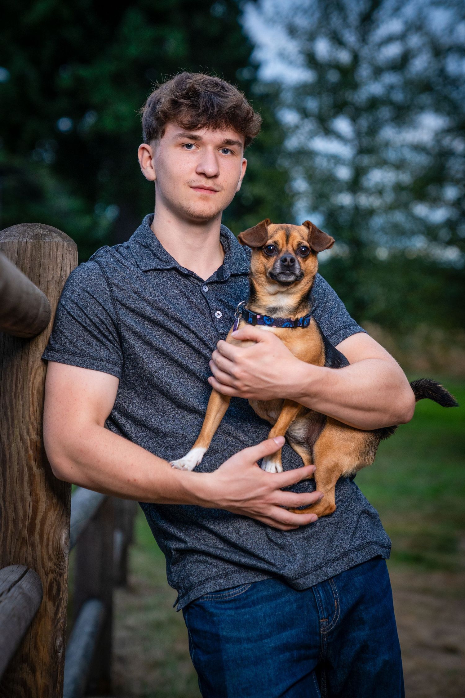 Person holds small brown dog while posing against wooden fence.