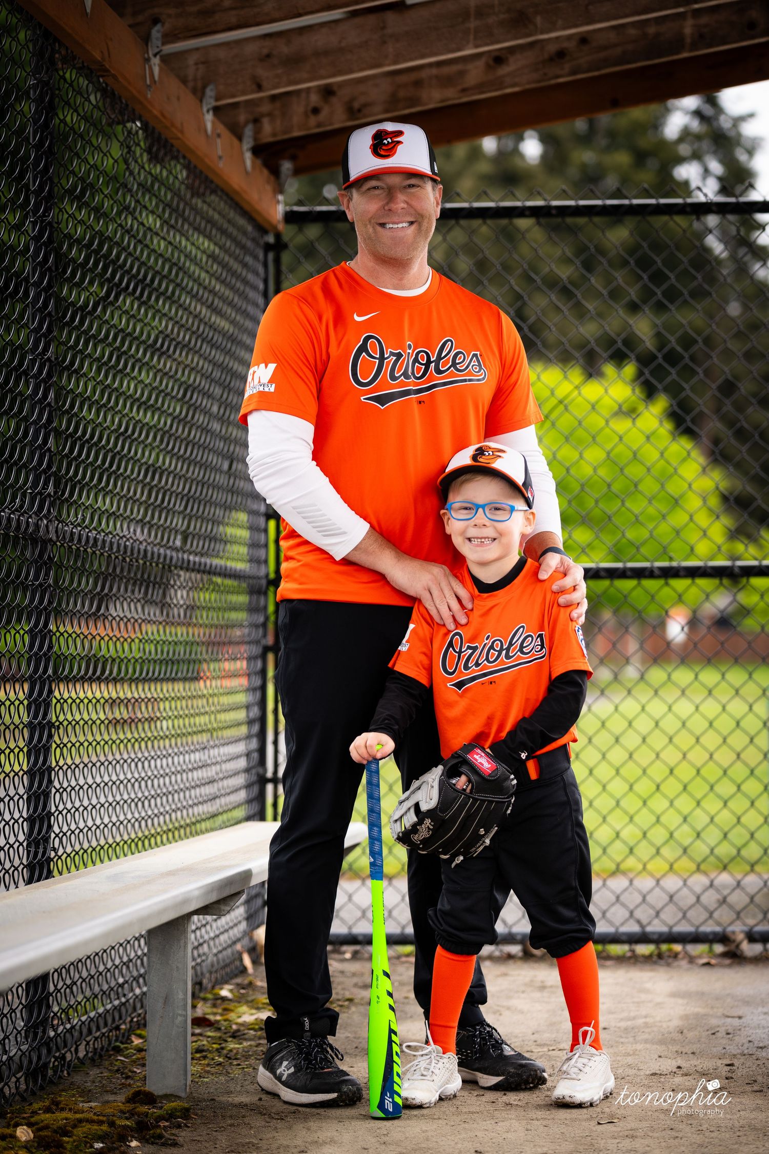 Two baseball players in orange Orioles uniforms stand together by a fence at a baseball field with bats and gear.