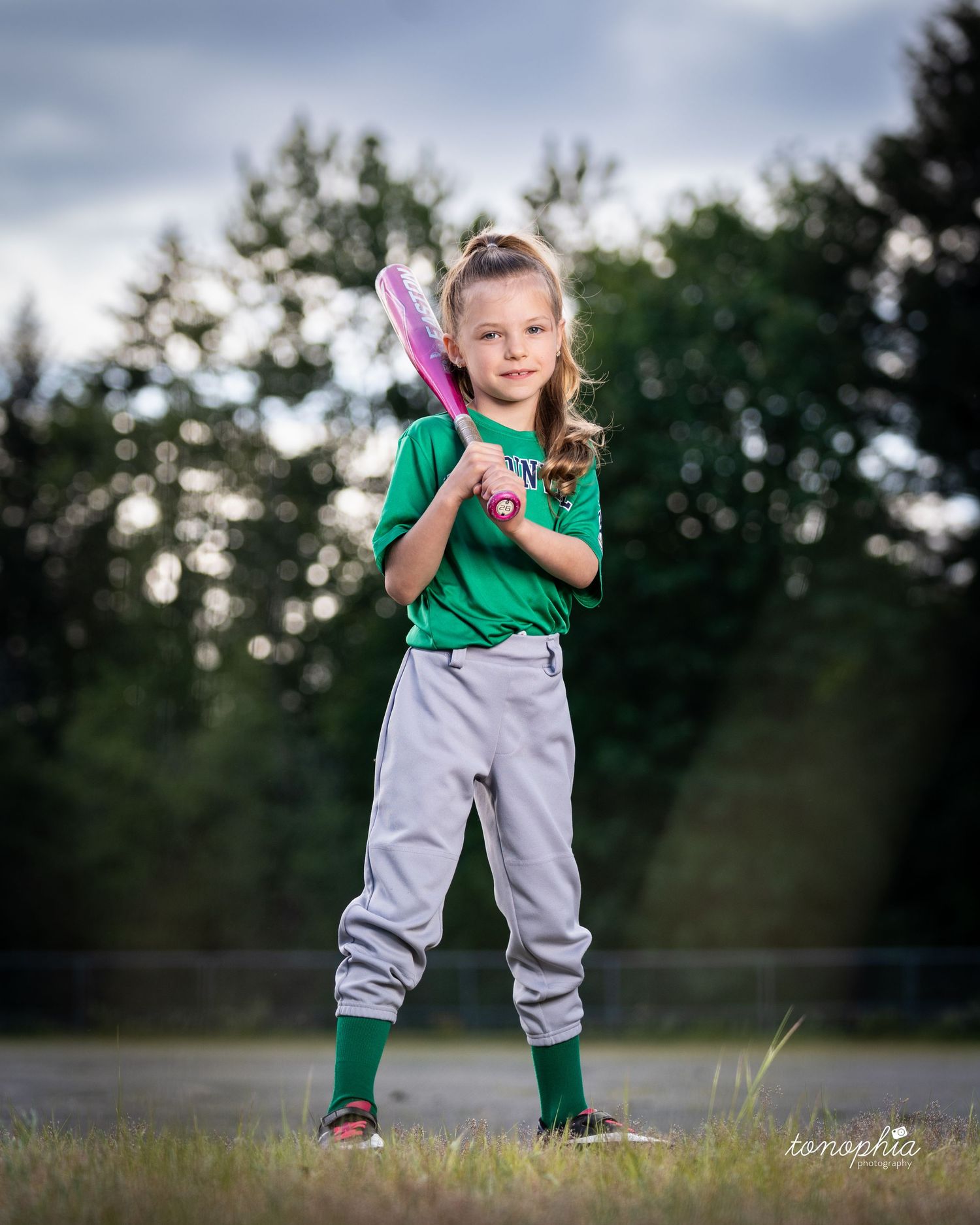 Baseball player in green and gray uniform stands ready with pink bat on outdoor field at dusk.