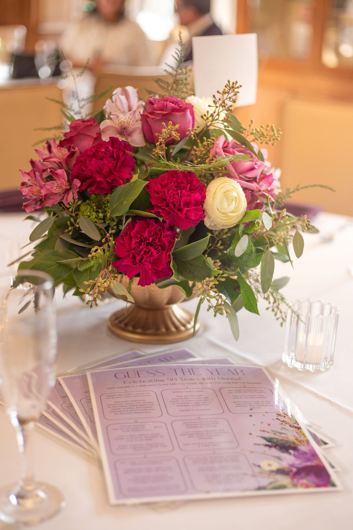 Elegant floral centerpiece with pink and red roses in gold vase next to menu cards on white tablecloth at event setting.