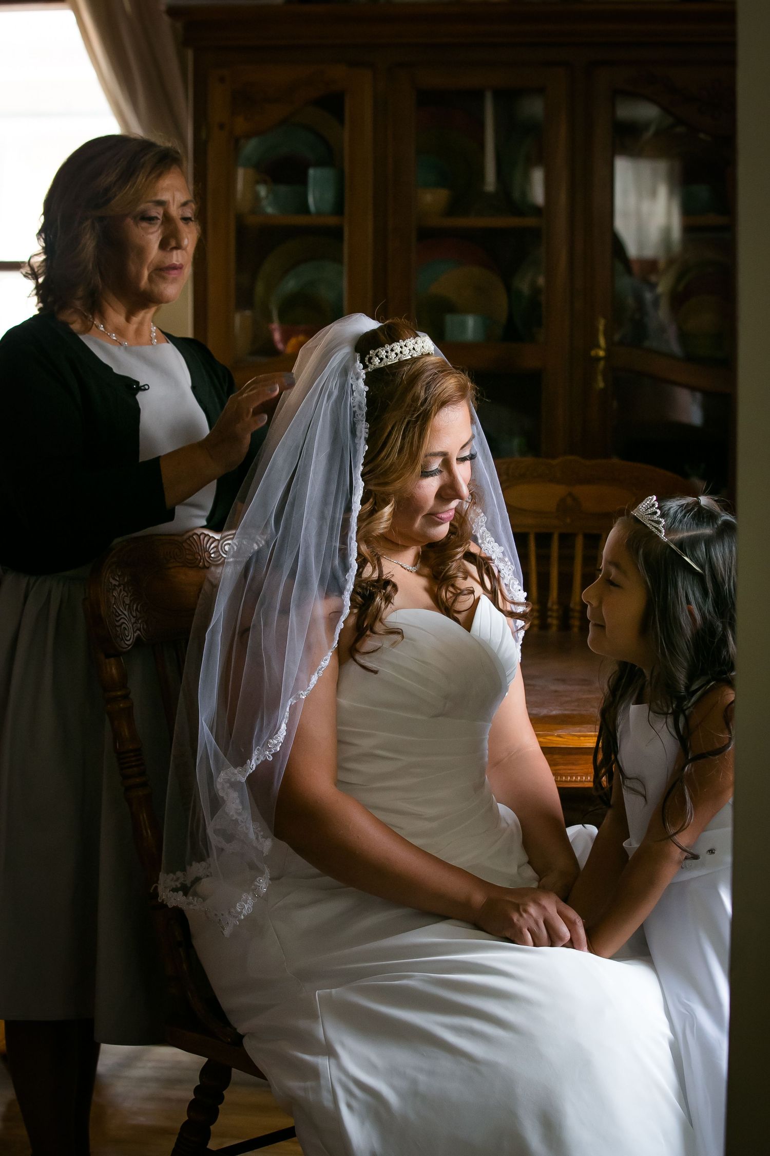 A bride in a white wedding dress sits in a wooden chair while being helped with final preparations in a dimly lit room.