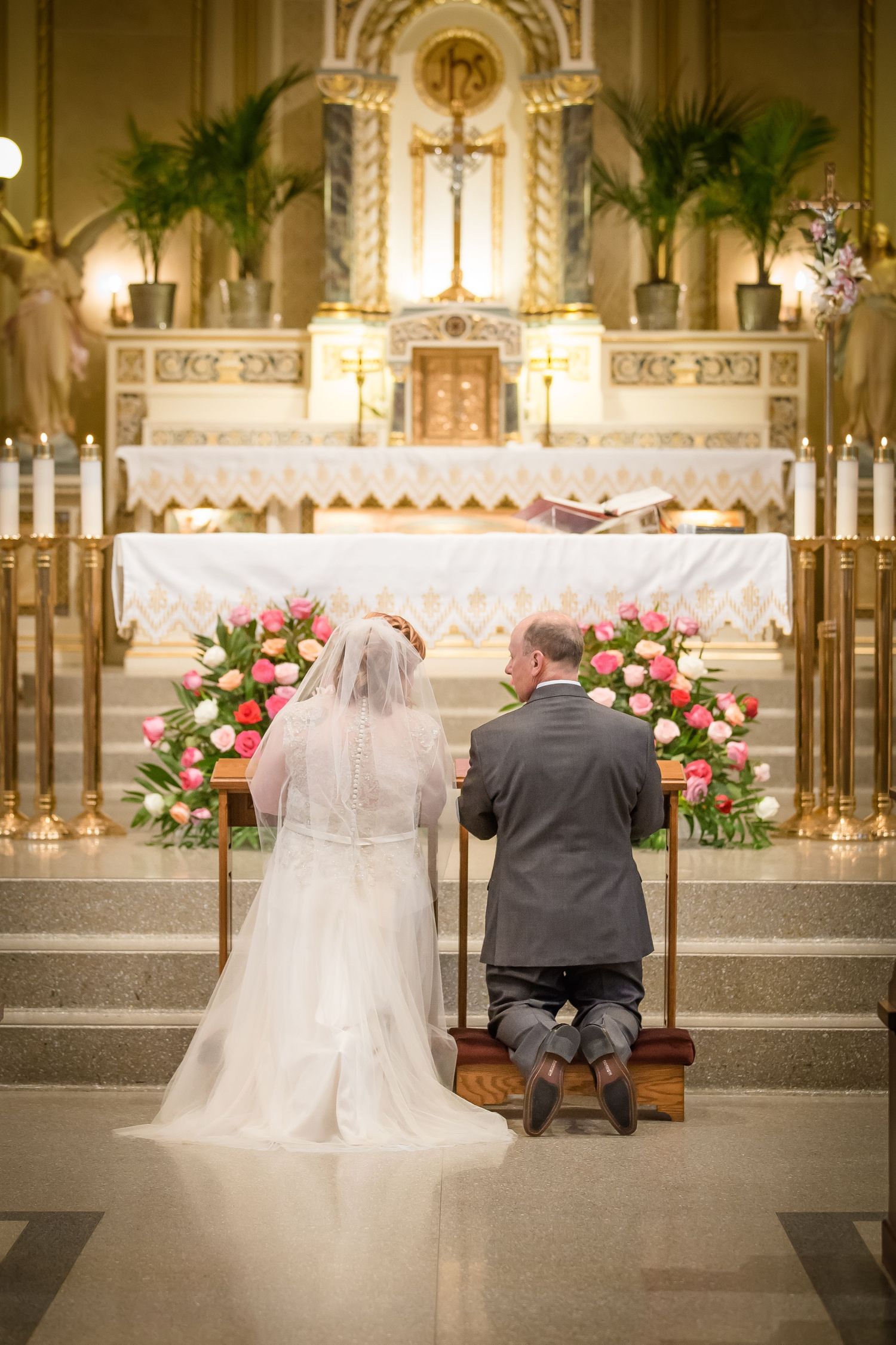 A couple sits together on a church pew in front of an ornate altar decorated with pink roses and greenery.