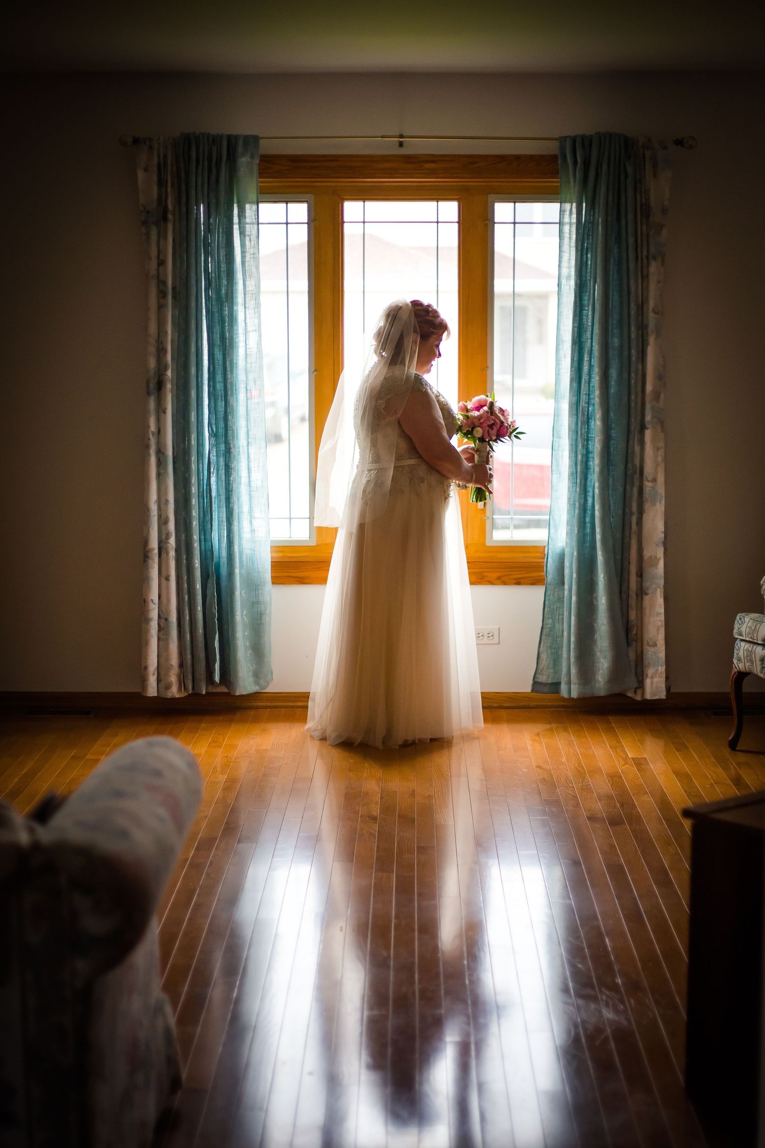 A wedding dress silhouetted against sunlit windows in a room with hardwood floors and turquoise curtains.