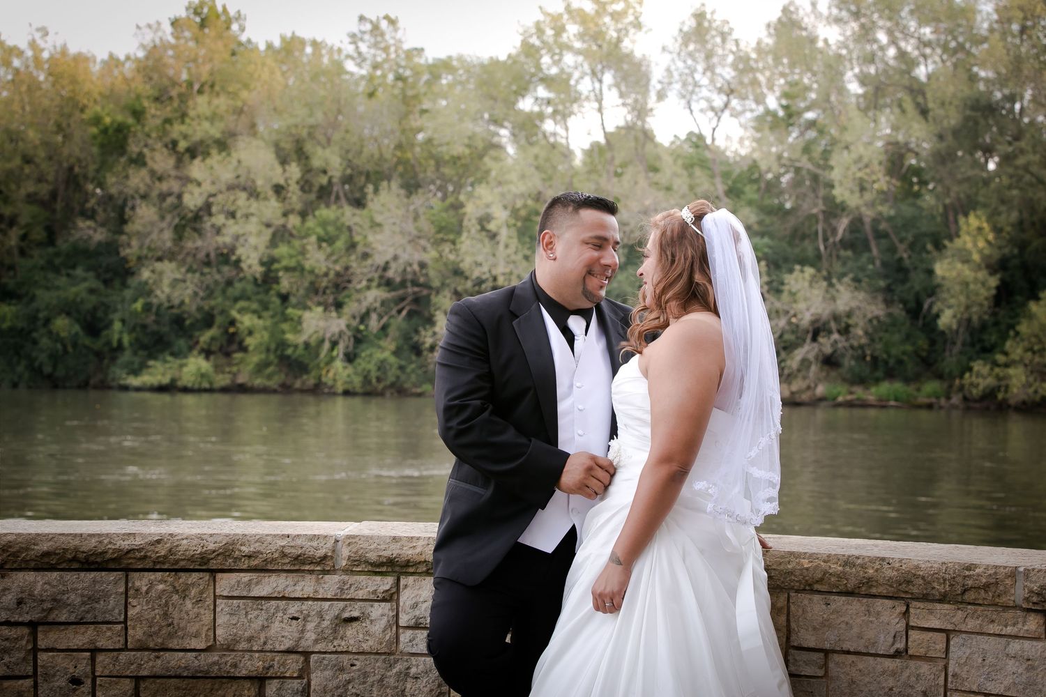 Newlywed couple shares romantic moments in a scenic outdoor setting with trees and a lake in the background.