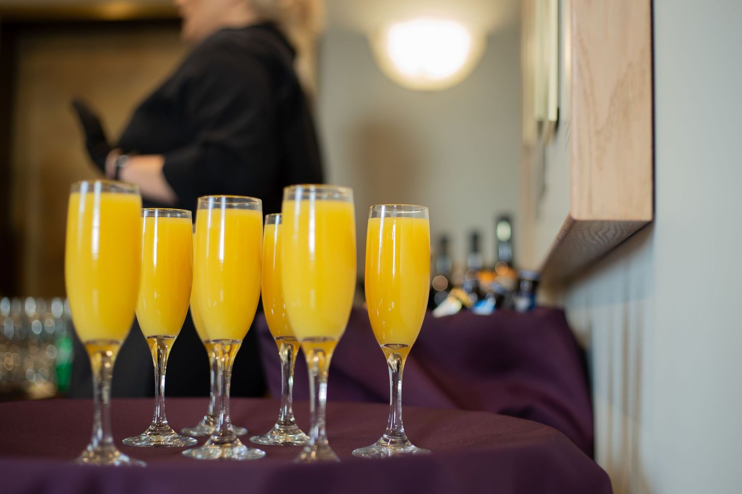 Five champagne flutes filled with bright orange mimosa cocktails lined up on a purple tablecloth.