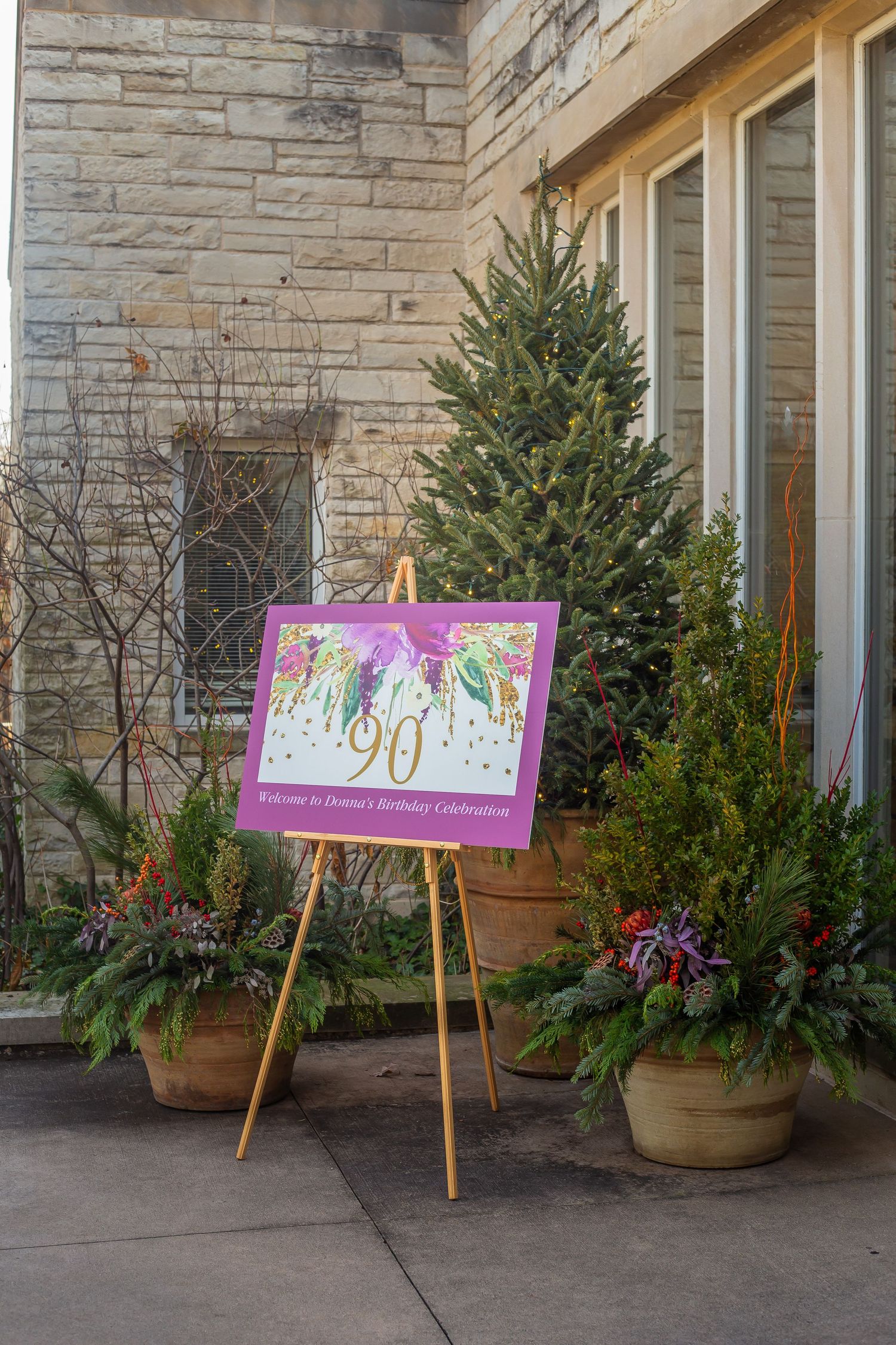 A series of outdoor holiday displays with evergreen trees and decorative signs on wooden easels against a stone wall.