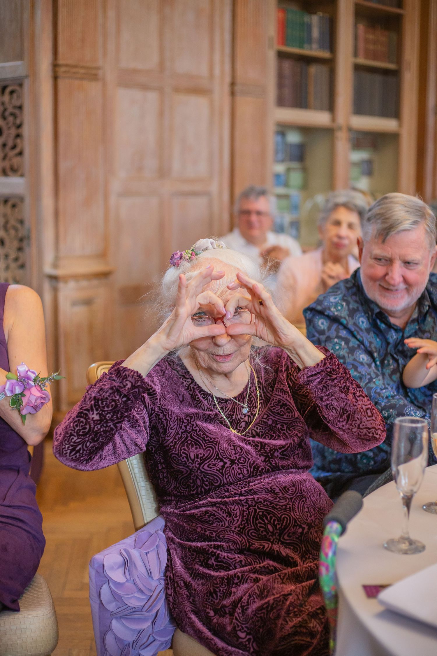 A person in a burgundy velvet top makes heart shapes with their hands while seated at a table.