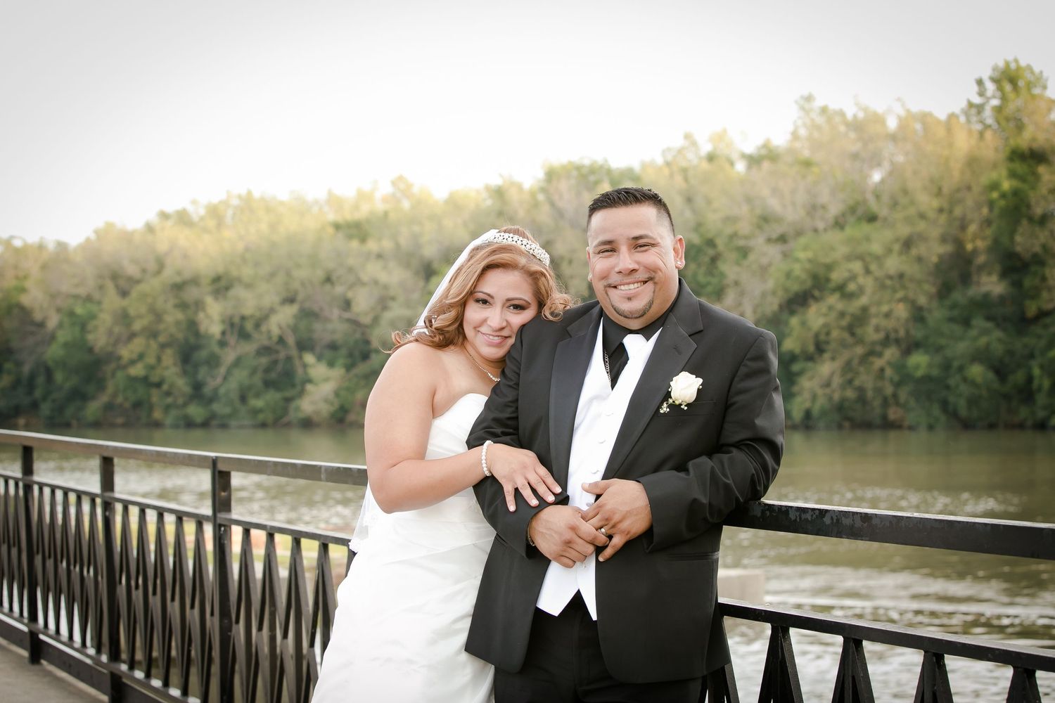A happy newlywed couple shares a joyful moment on a bridge railing overlooking a serene river at sunset.
