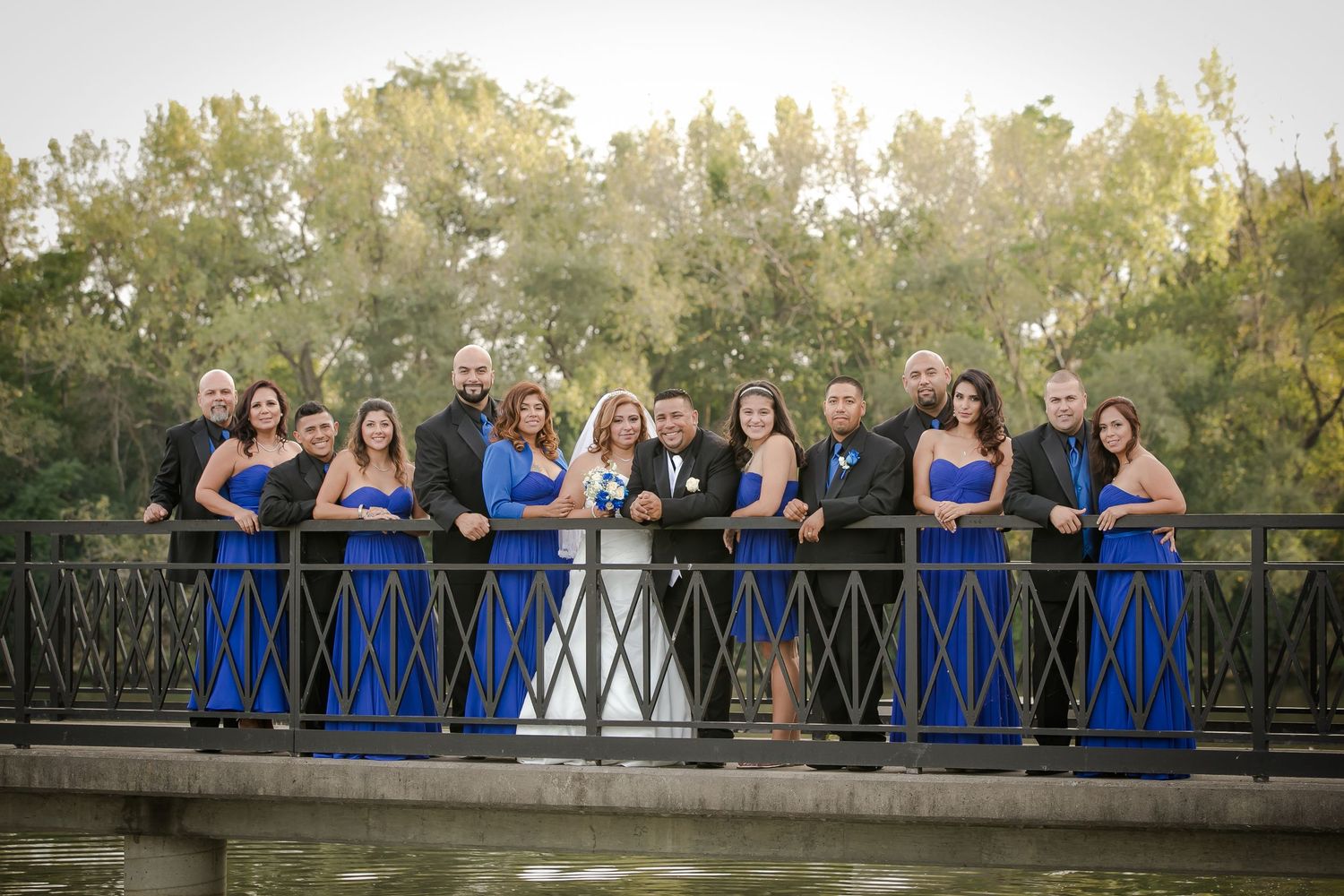 Wedding party in coordinating blue and white dresses pose together on a rustic bridge in an outdoor setting.