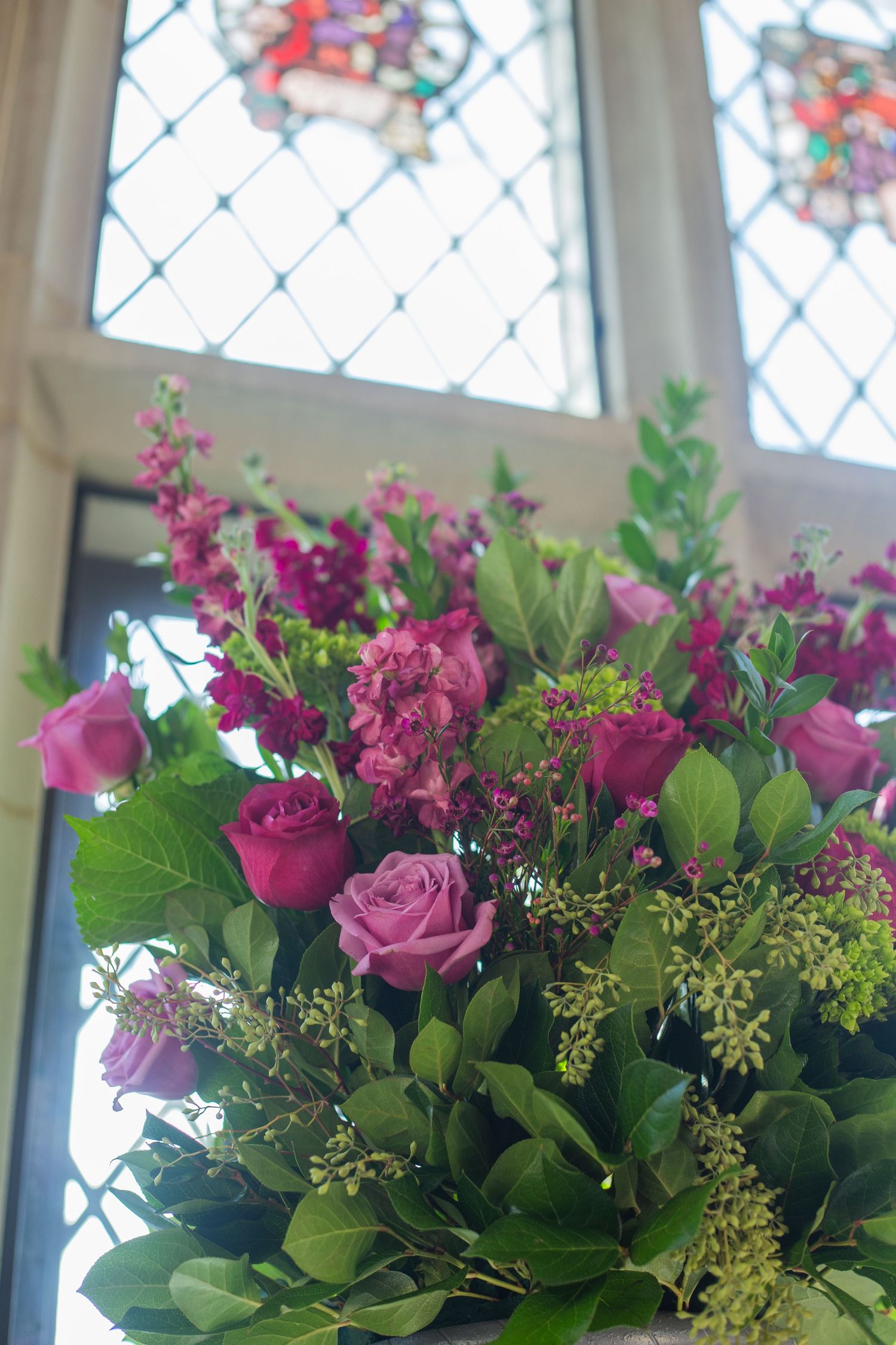 Beautiful pink roses and floral arrangement with delicate greenery displayed against a stained glass church window background.