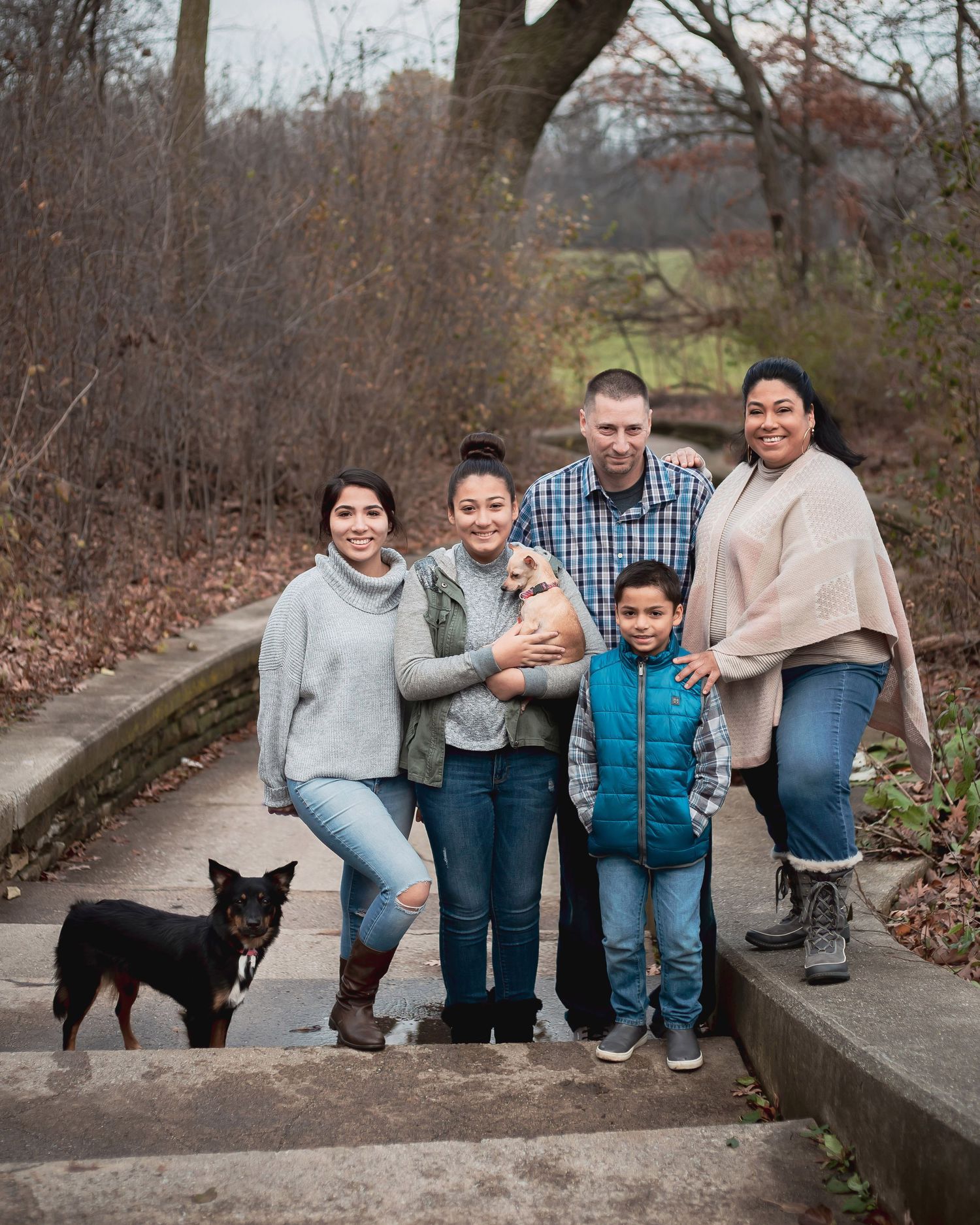 Family of five posing outdoors on stone steps in autumn park with two dogs, smiling for a portrait photo.