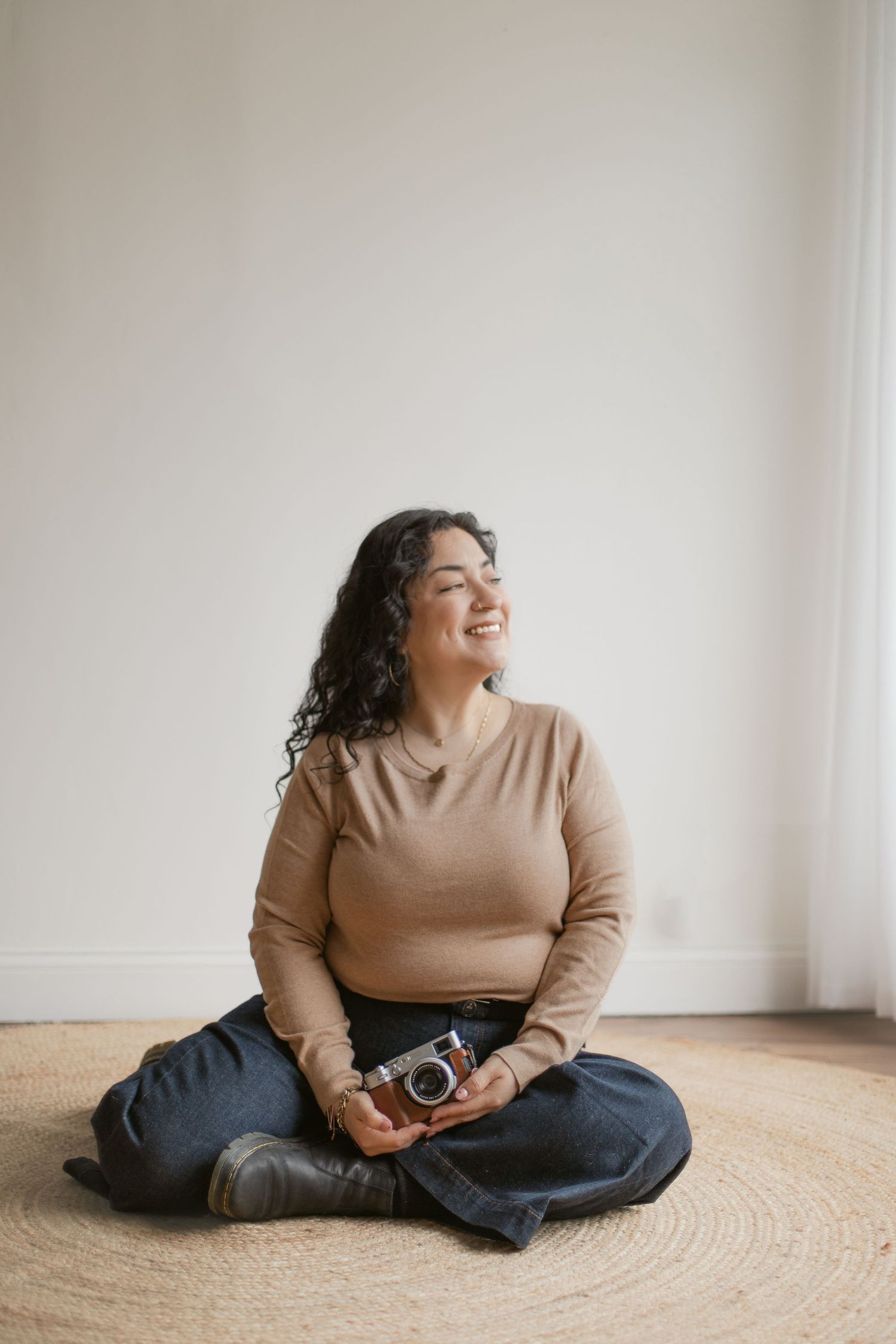 Smiling photographer in beige sweater and jeans sits cross-legged on round rug while holding vintage camera.