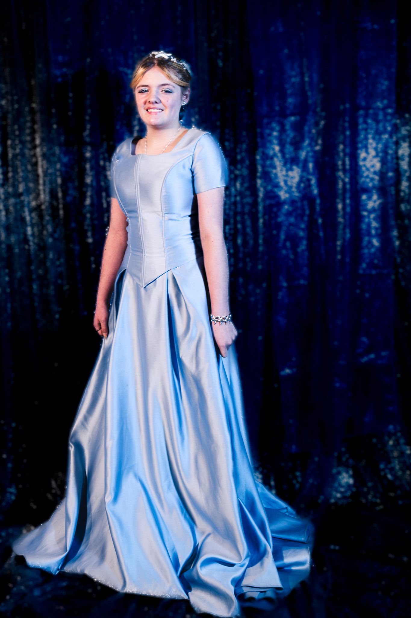 A model in a flowing ice blue satin gown walks the runway against a dark blue backdrop.