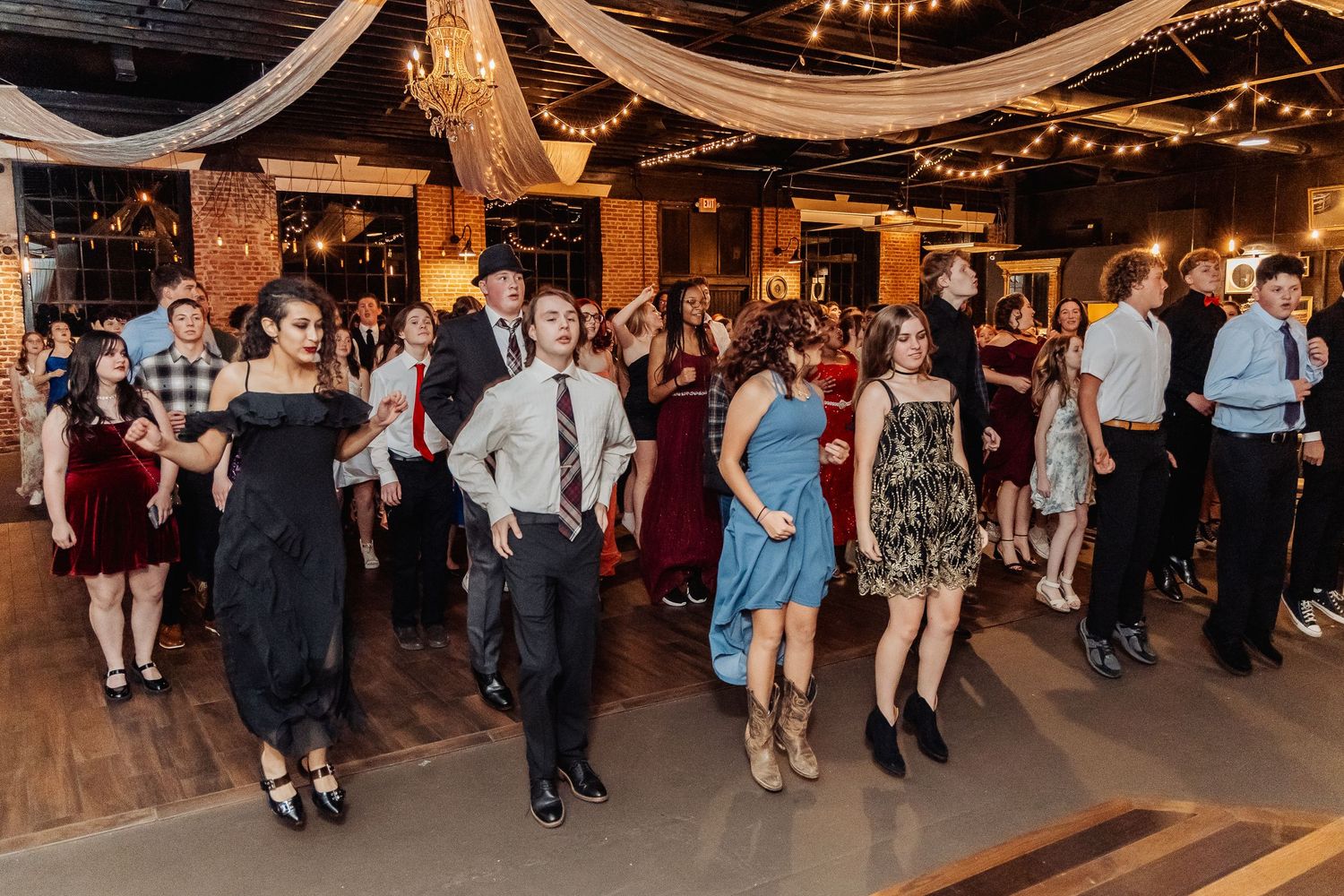 People dancing and socializing at an indoor event venue decorated with string lights and white fabric draping.