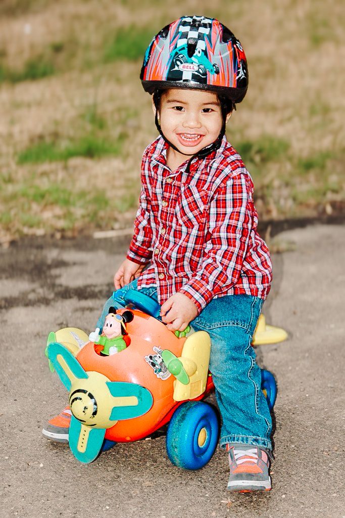 A child wearing a plaid shirt and safety helmet plays with a colorful orange toy airplane on pavement.