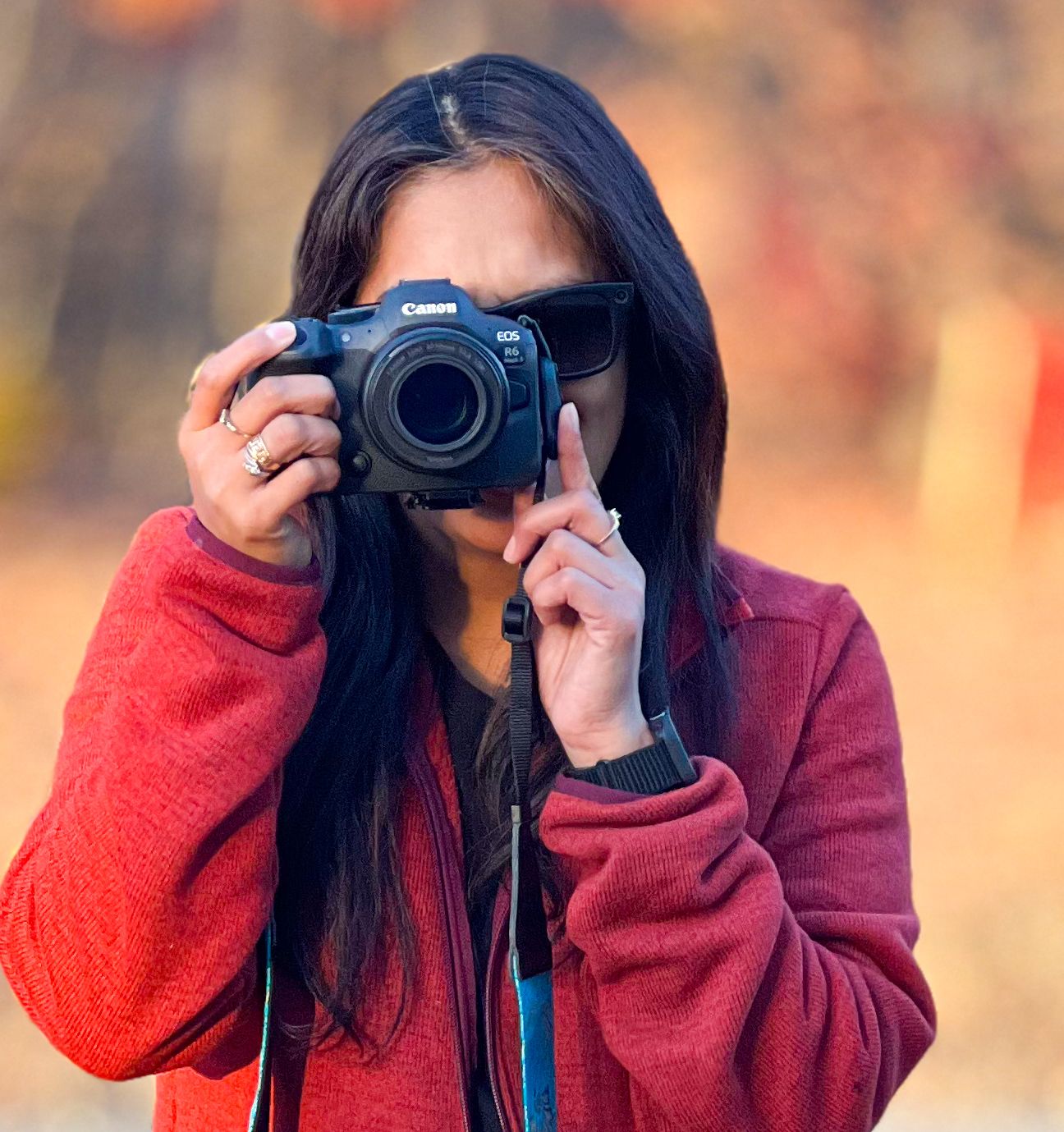 Person in red sweater taking photos with DSLR camera against blurred autumn background.
