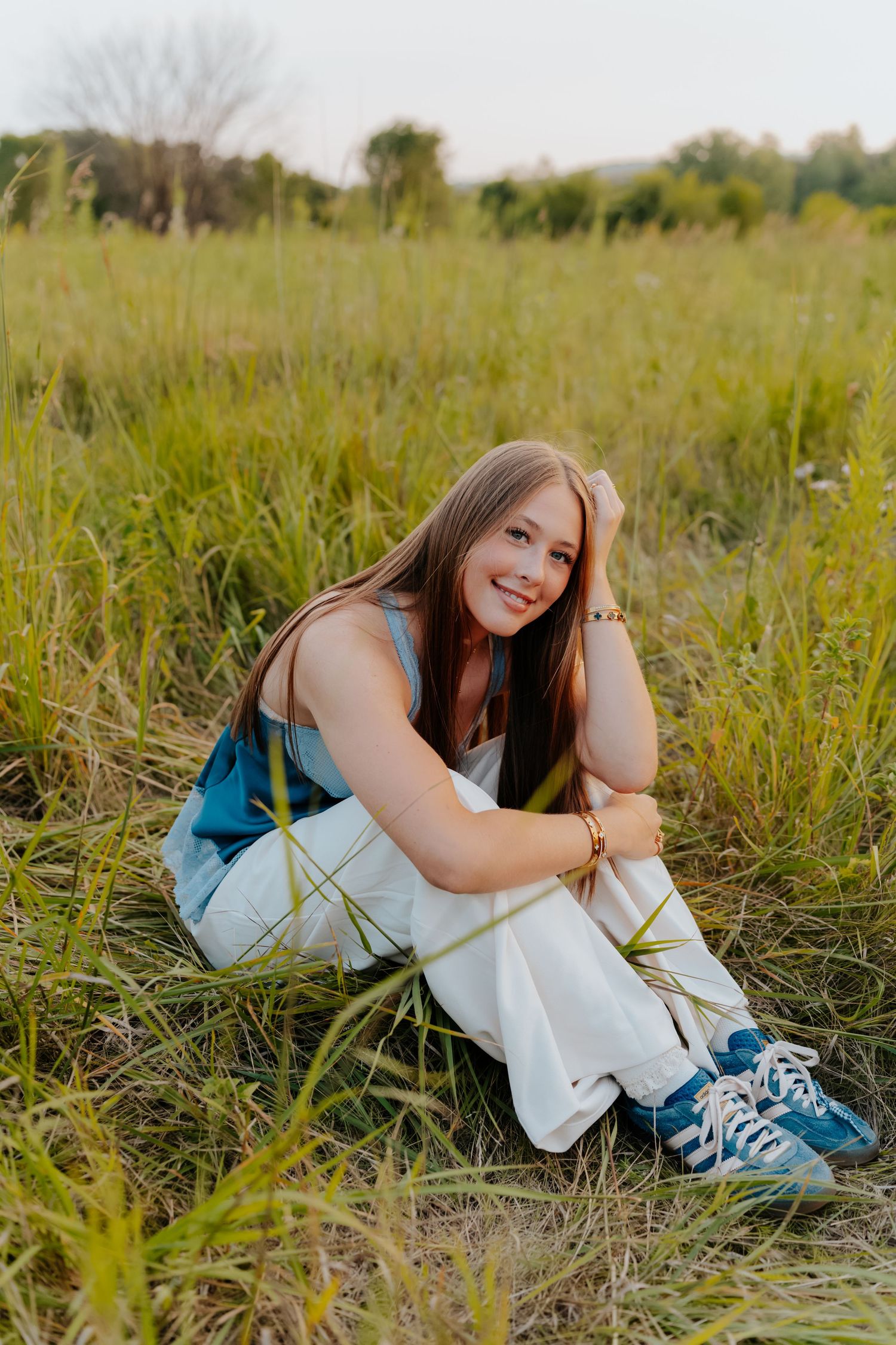 Young woman in white dress and blue sneakers sitting in a lush green field, smiling warmly at the camera.