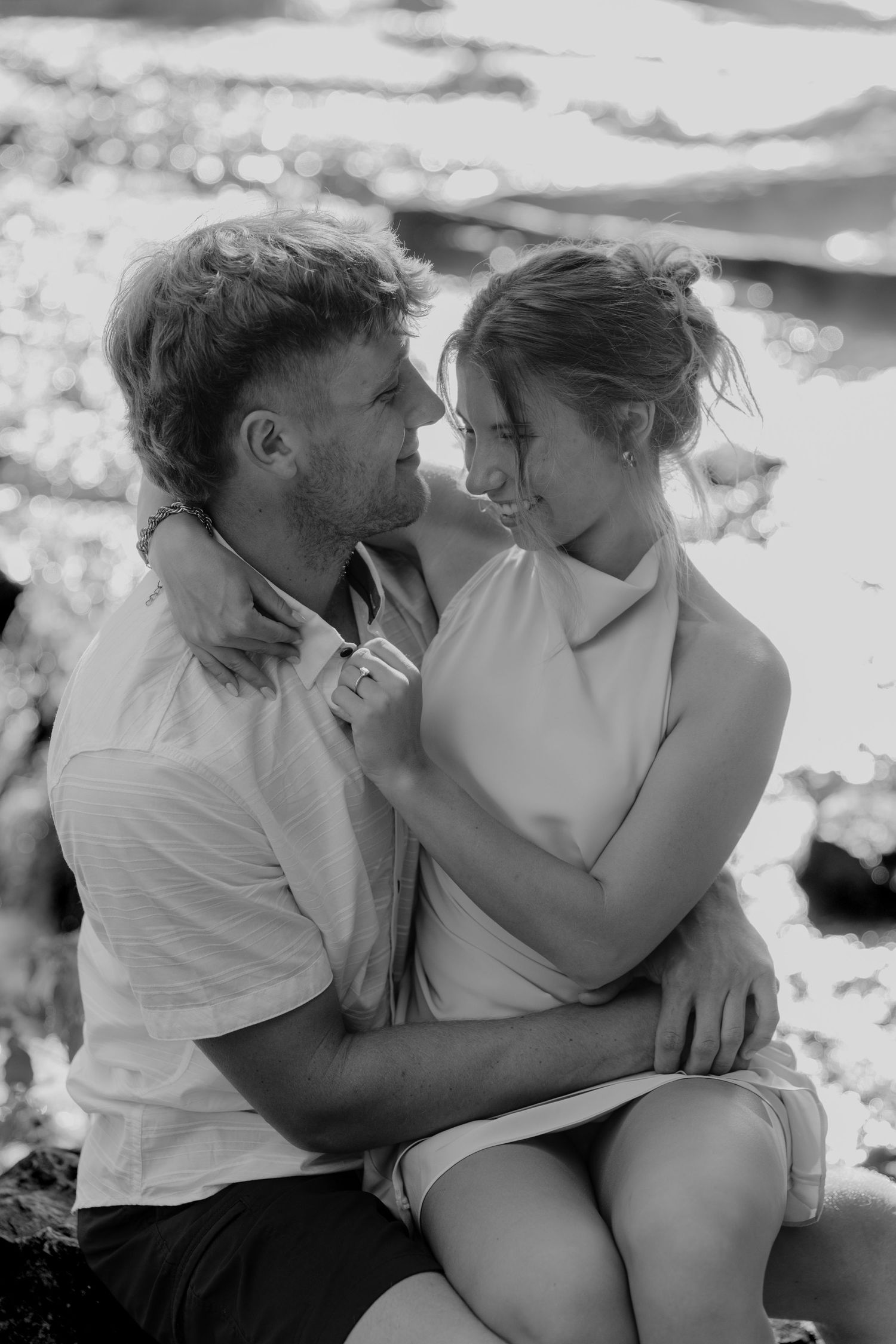 Black and white photo of a romantic couple embracing and gazing at each other outdoors in soft natural light.