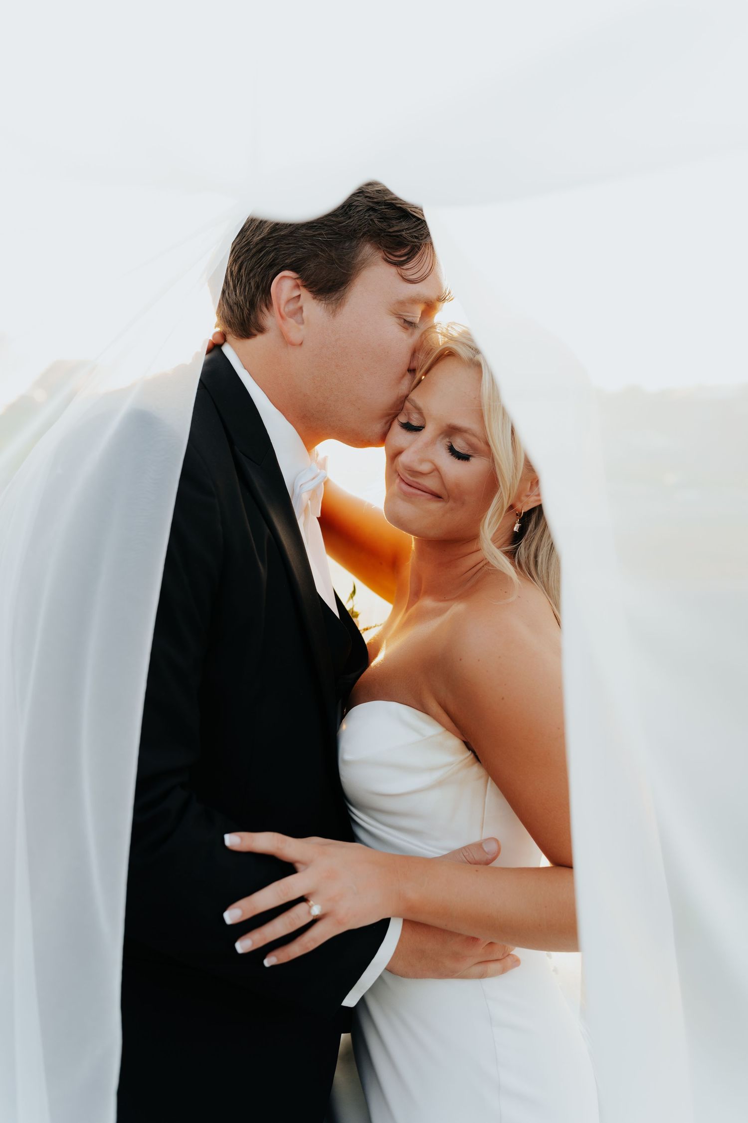 Groom in black tuxedo kissing bride on forehead, wrapped in flowing white veil during golden hour.
