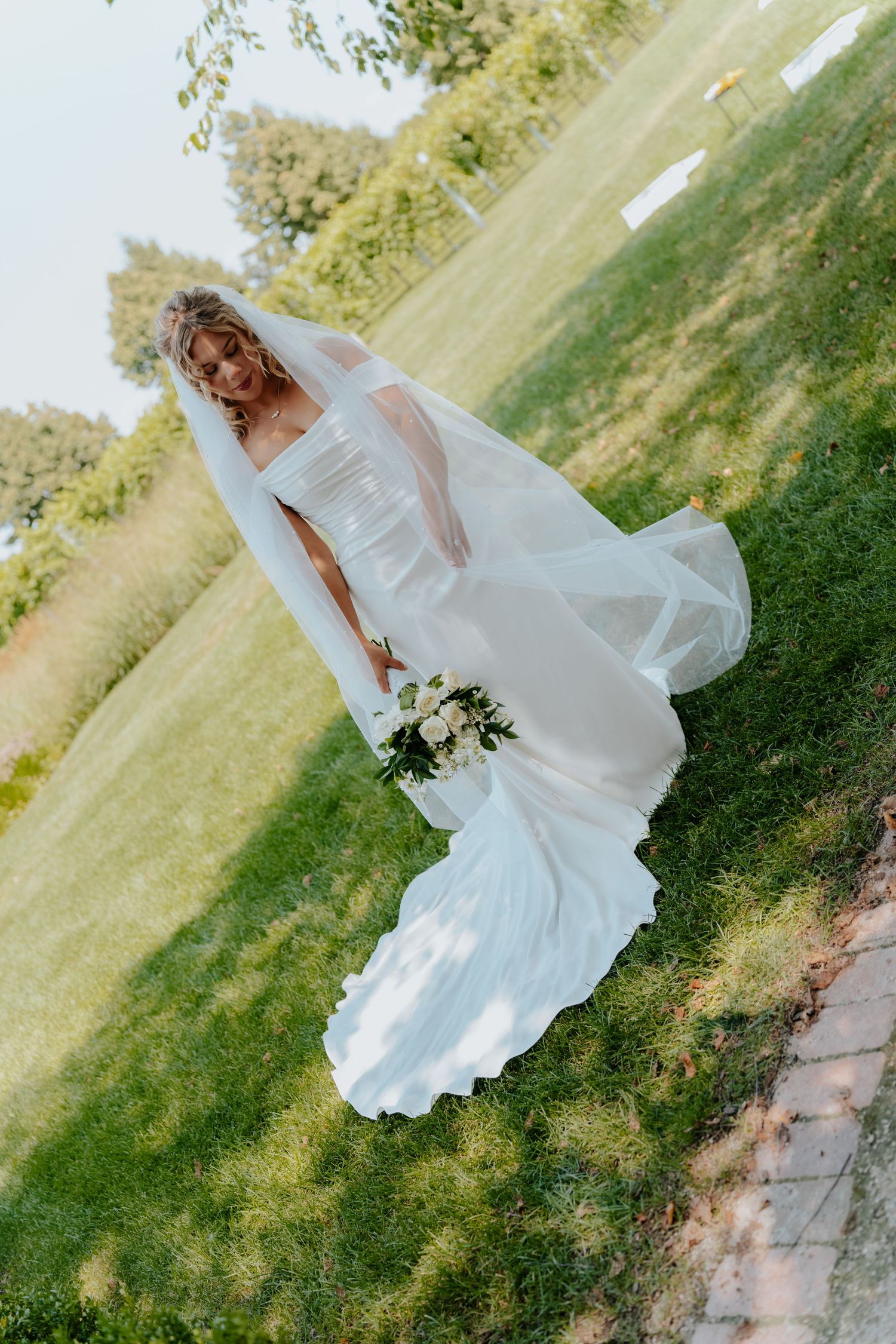 Bride in white gown and veil holds bouquet on green lawn.