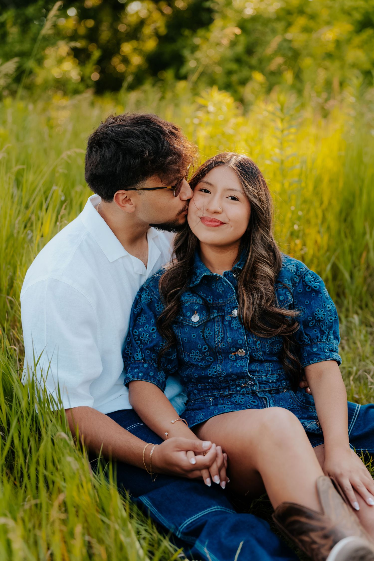 A couple sits in a lush green field, sharing a tender kiss on the cheek during a romantic outdoor engagement session.