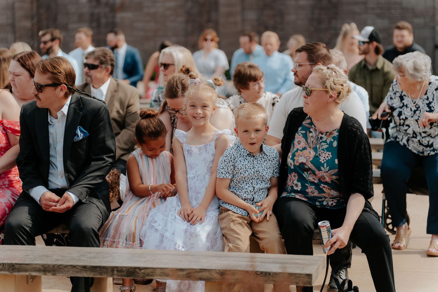 Wedding guests including children seated on benches during an outdoor wedding ceremony.