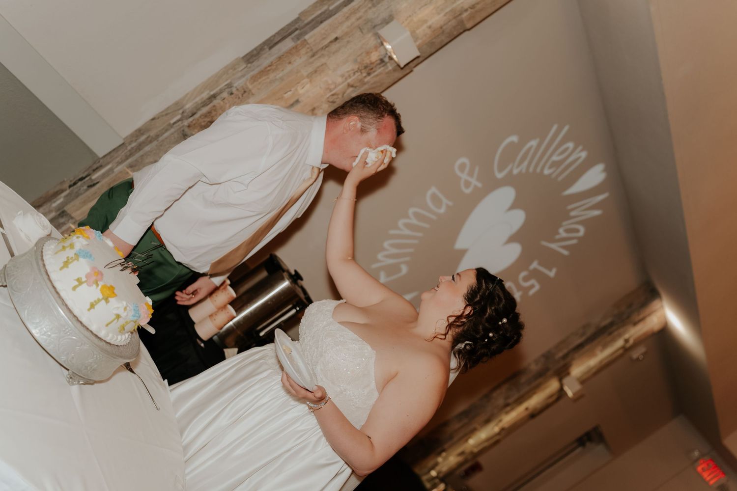 Bride smearing wedding cake on groom's face during fun cake cutting ceremony at elegant reception venue.