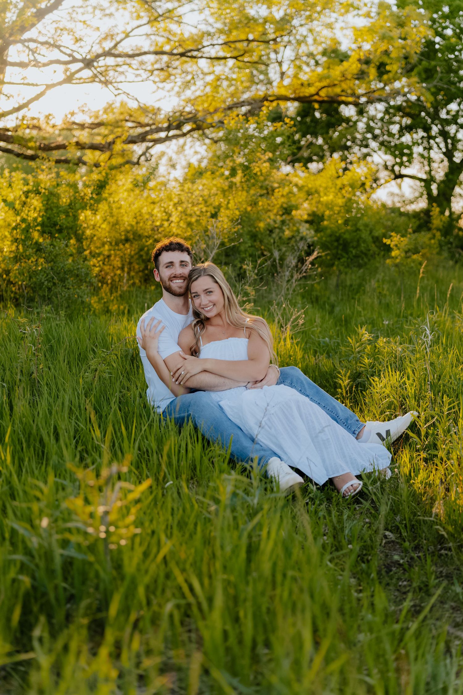 Happy couple sitting in lush green meadow at golden hour, man embracing woman in white dress and jeans.