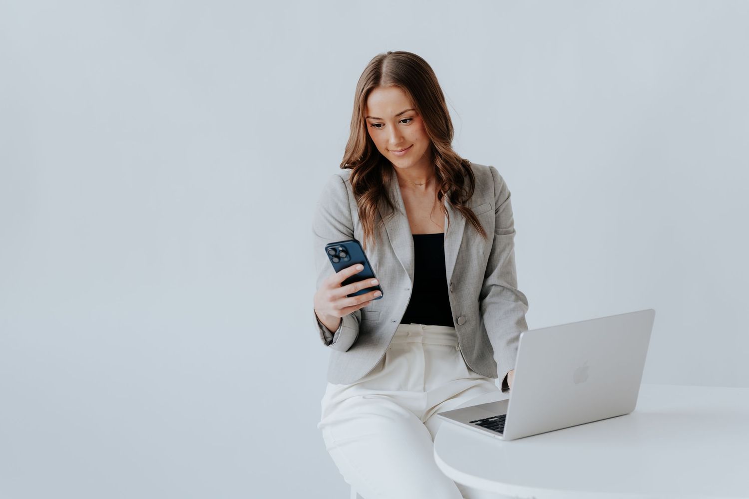 Professional woman in blazer smiling at smartphone while sitting at desk with open laptop on white background.