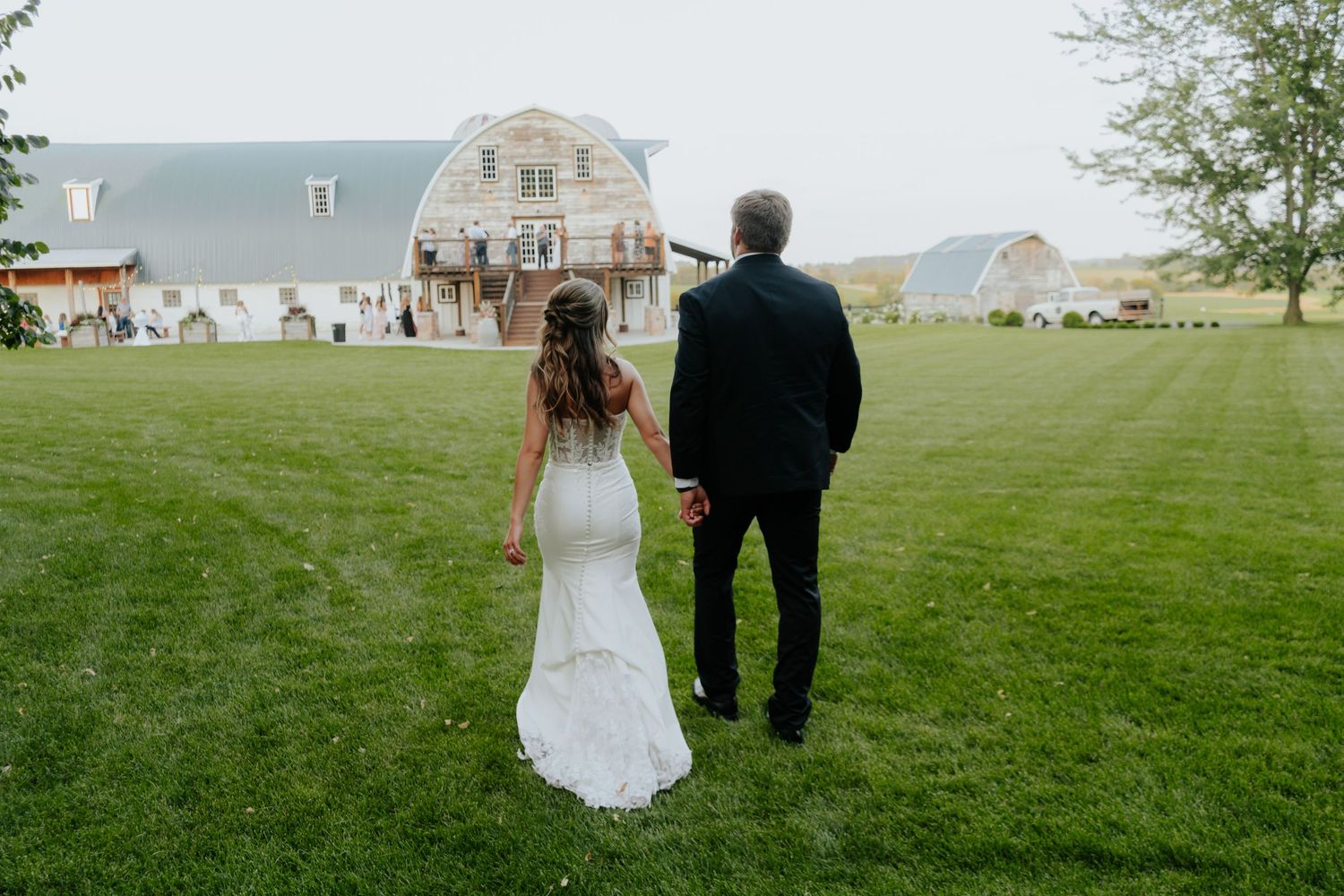 A couple walks hand in hand across a lush green lawn with a rustic barn and farmhouse in the background.