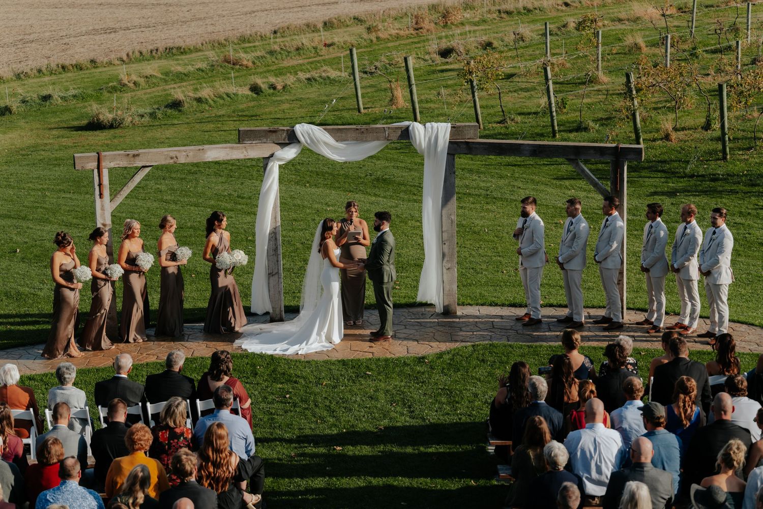 Outdoor wedding ceremony under a rustic wooden arch with white draping on a lush green hillside.