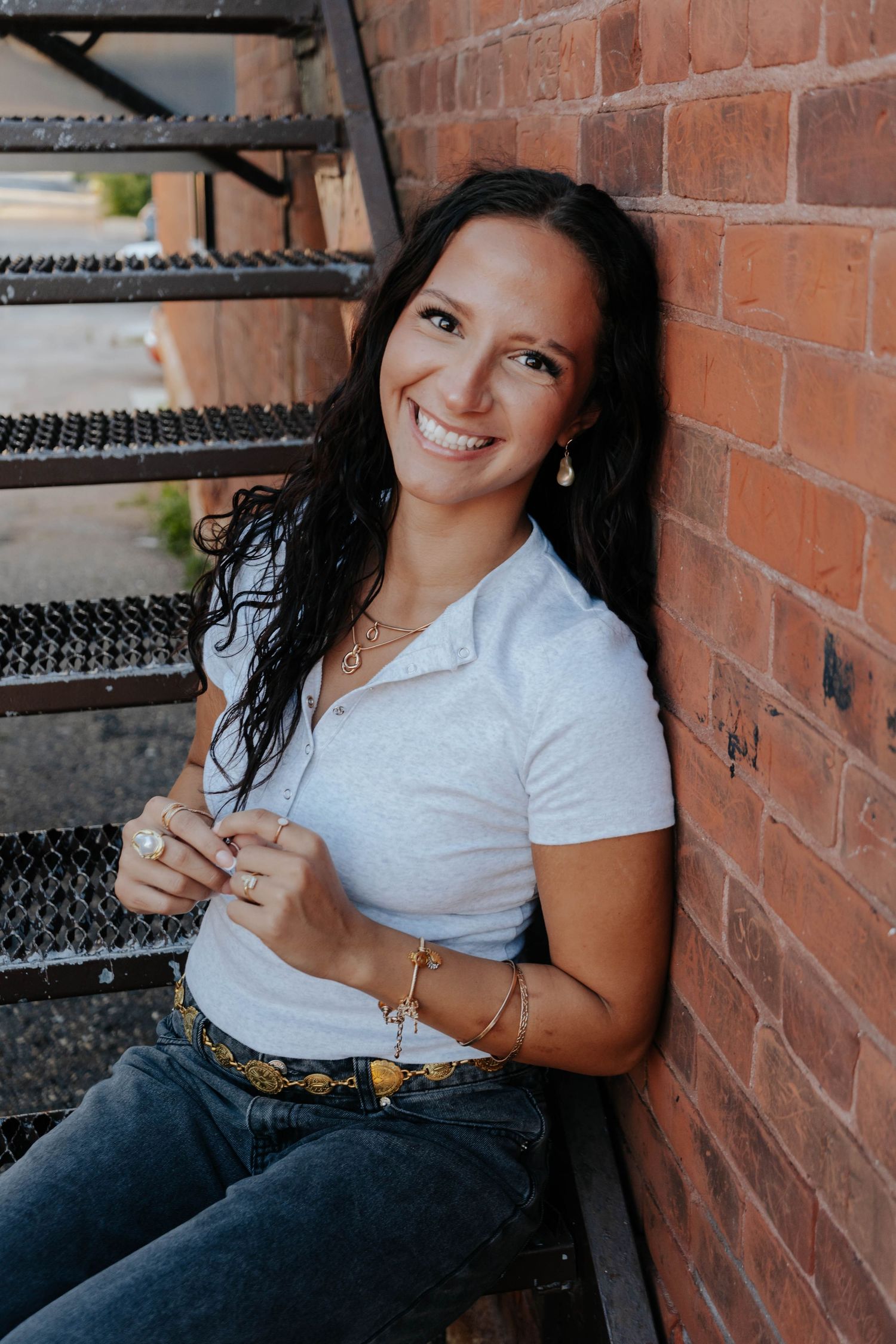 Smiling young woman with dark curly hair in white polo and jeans, leaning against brick wall on metal stairs.