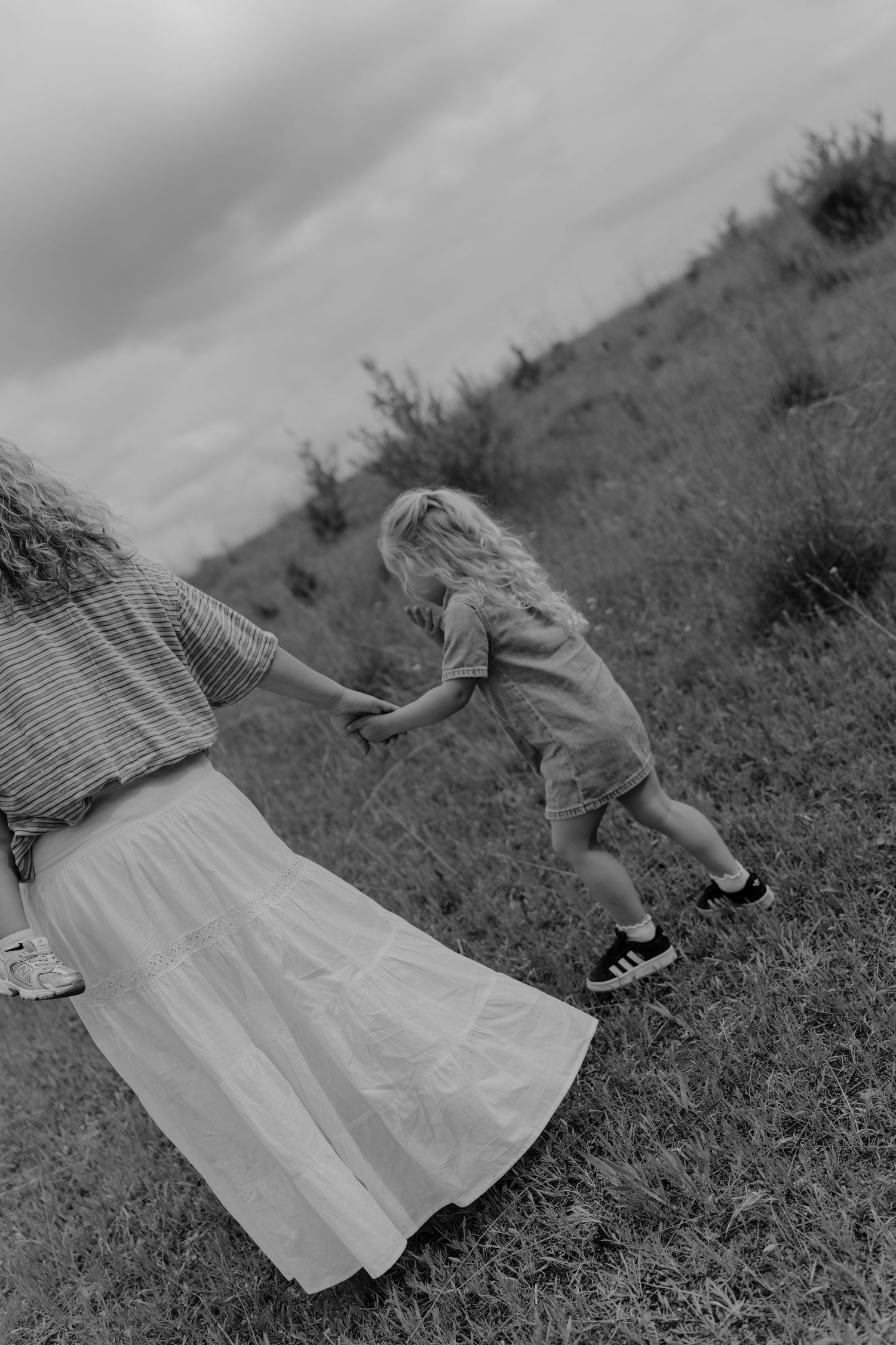 Black and white photo of a mother in a white skirt guiding a curly-haired child running on a grassy hillside.