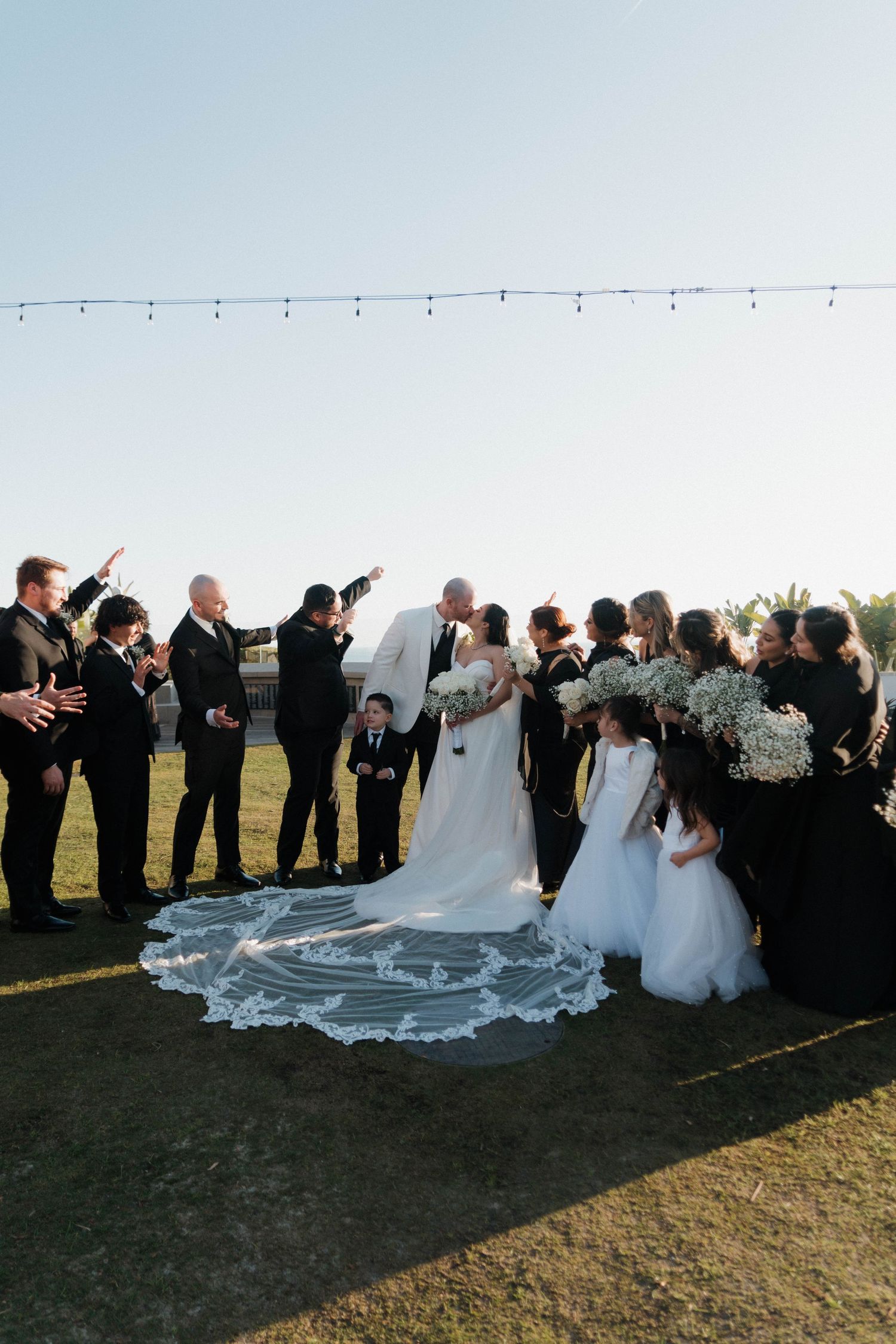 Wedding party gathered on grassy field at sunset with bride's long lace veil trailing behind during outdoor ceremony.