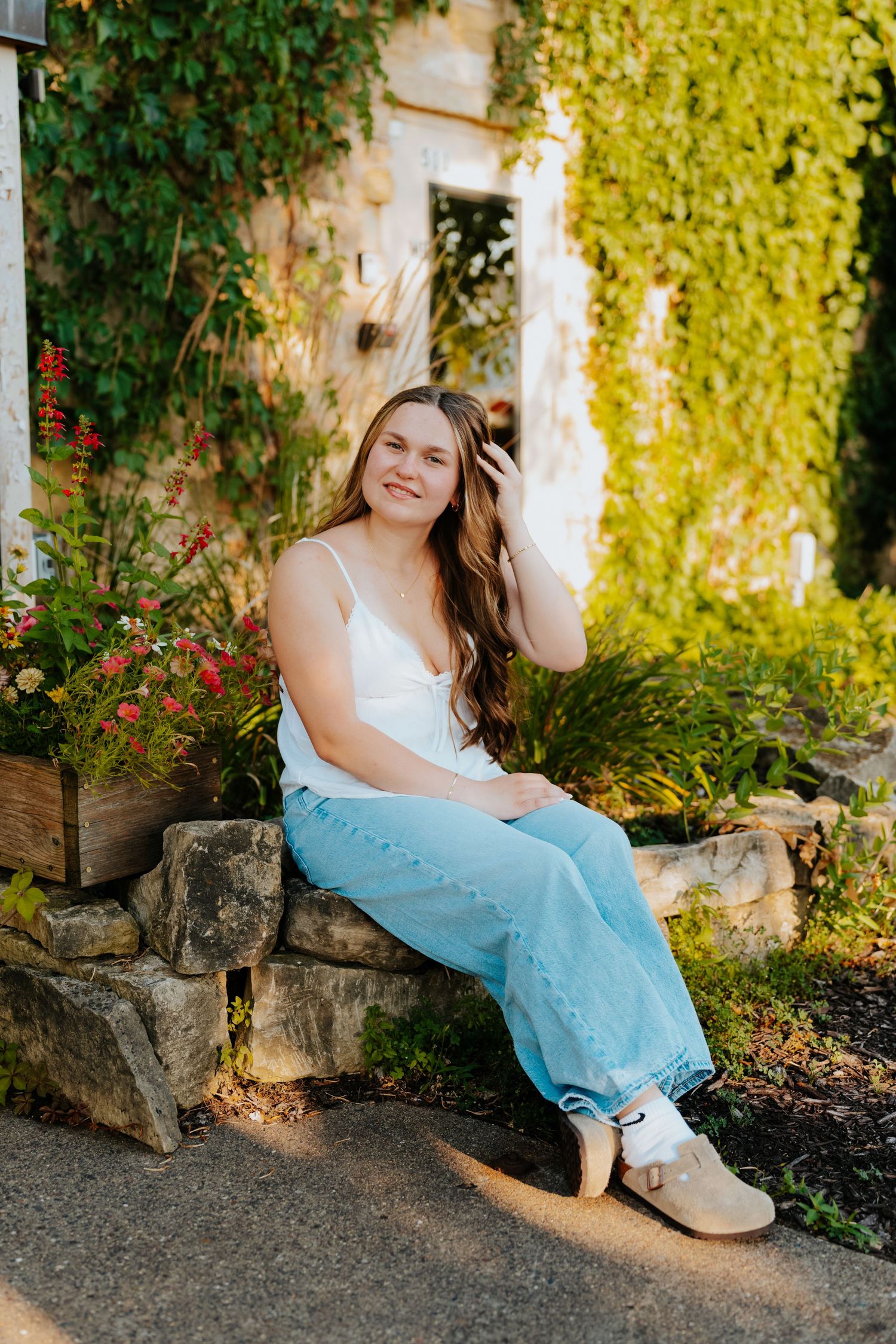 Young woman in white top and jeans sits on stone wall surrounded by lush green ivy and colorful flowers, smiling warmly.