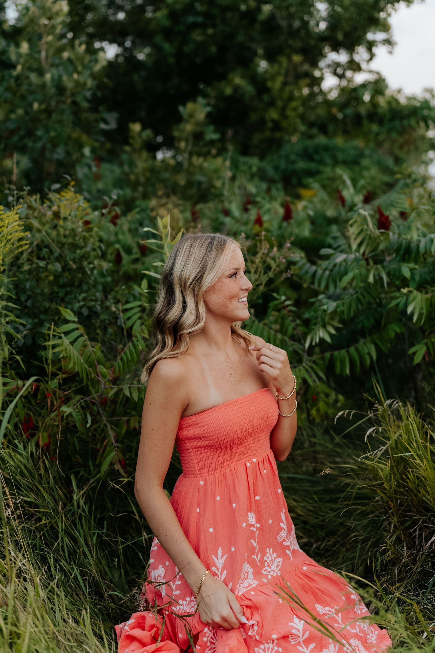 Woman in coral floral strapless dress smiling outdoors among lush green foliage and red flowers.