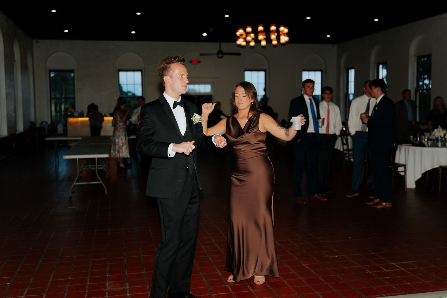 Couple dancing elegantly at a wedding reception, man in tuxedo and woman in brown gown, guests mingling in background.