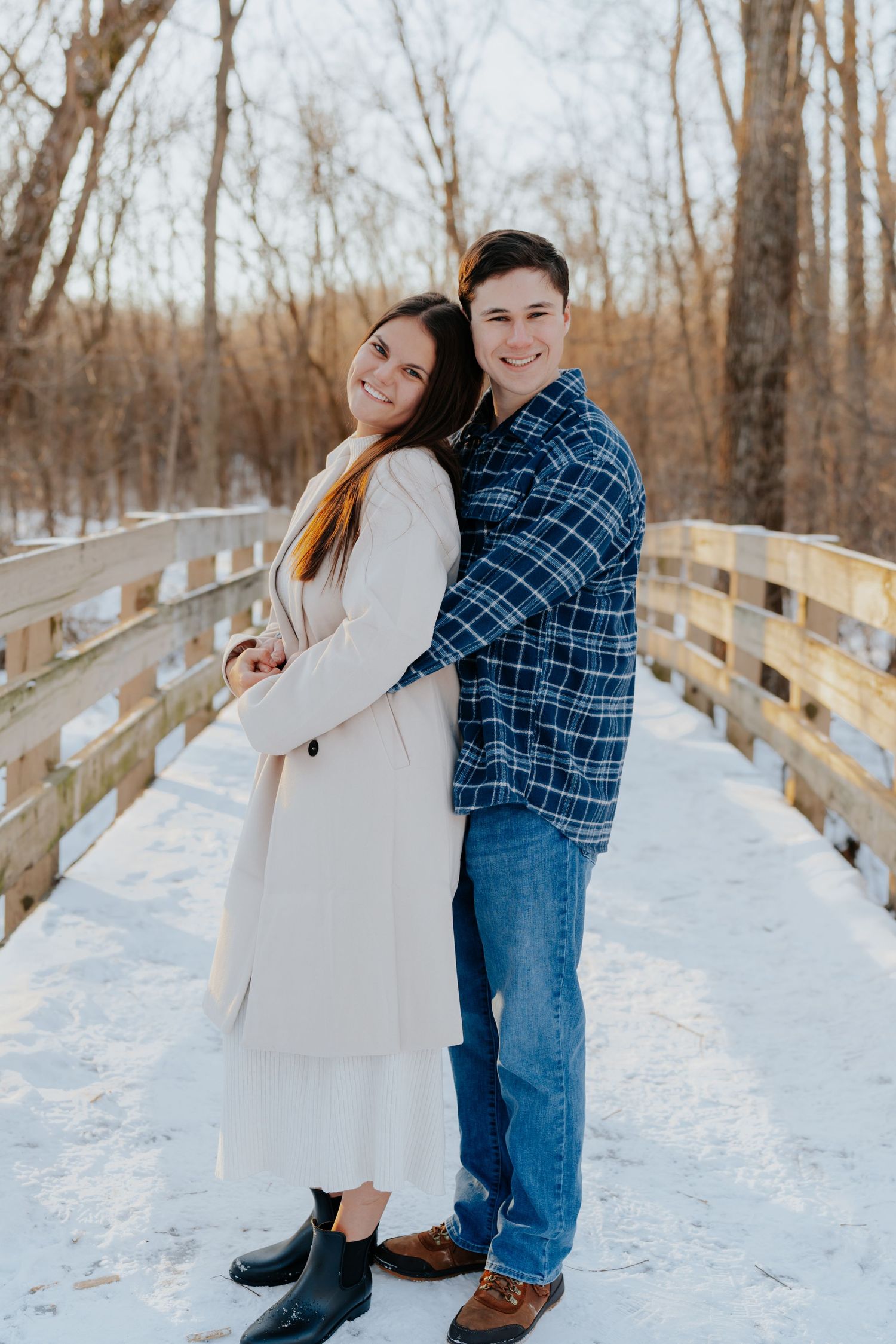 A couple poses back-to-back on a snowy wooden bridge, smiling, wearing a white coat and blue plaid shirt.