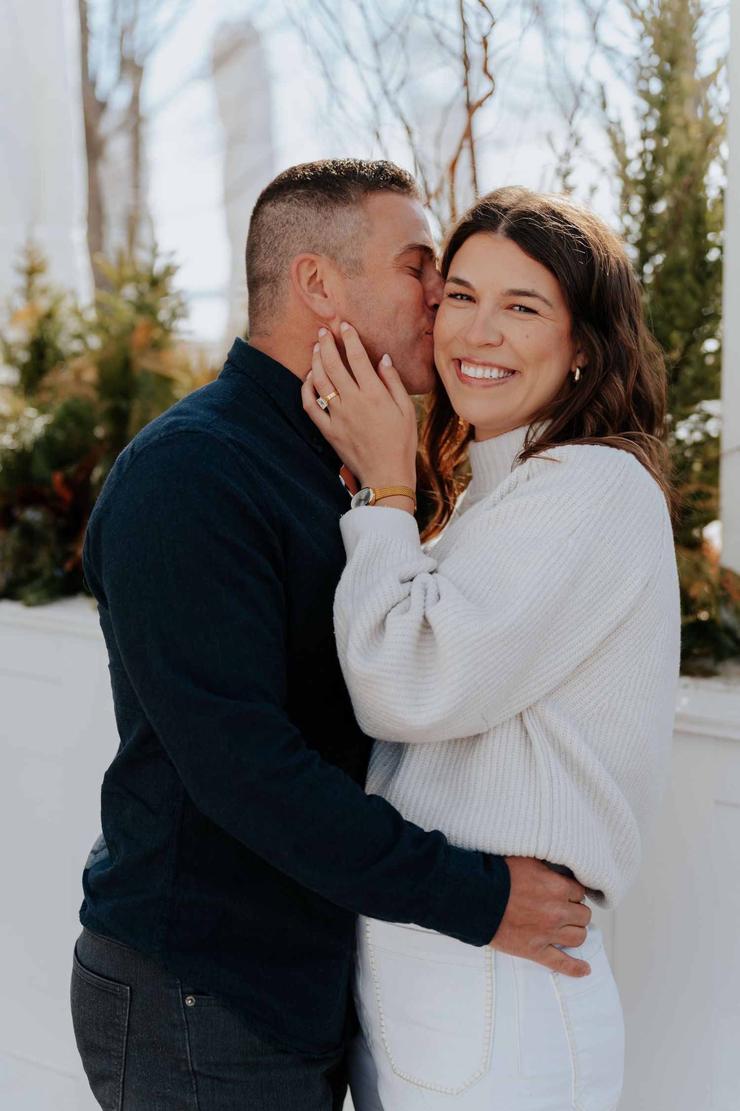 A couple shares a romantic moment, man kissing woman's cheek as she smiles, both wearing cozy sweaters outdoors.
